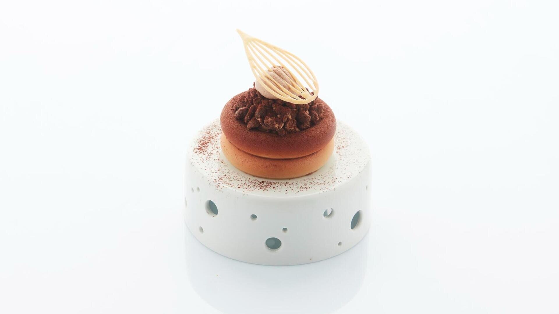 Ein kleines, elegantes Dessert mit rundem Boden, Schokoladenüberzug und einer zarten Verzierung, präsentiert auf einem weißen, durchbrochenen Sockel vor weißem Hintergrund.