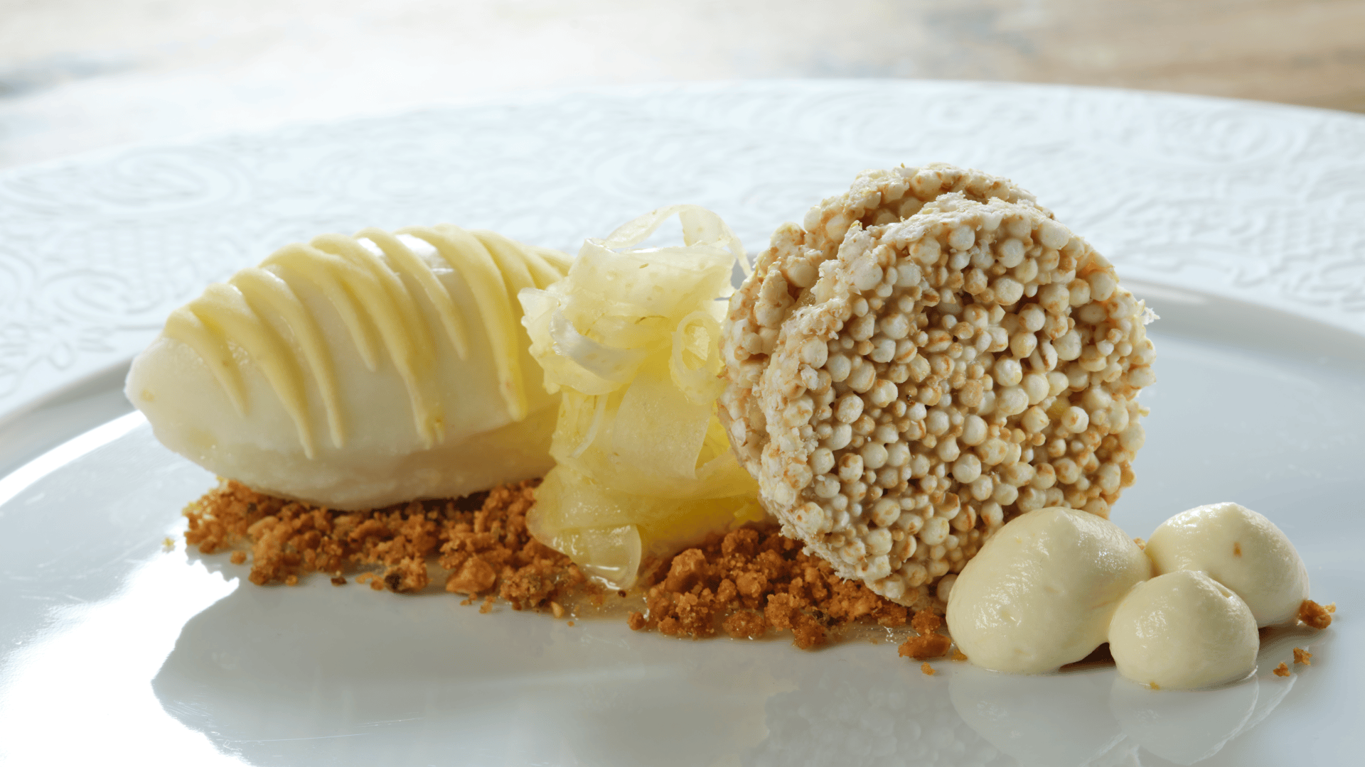 Ein auf einem Teller angerichtetes Dessert mit einer Kugel Sorbet, Fenchelscheiben, einer Scheibe aus gepufftem Getreide, Kekskrümeln und drei Tupfen Sahne.