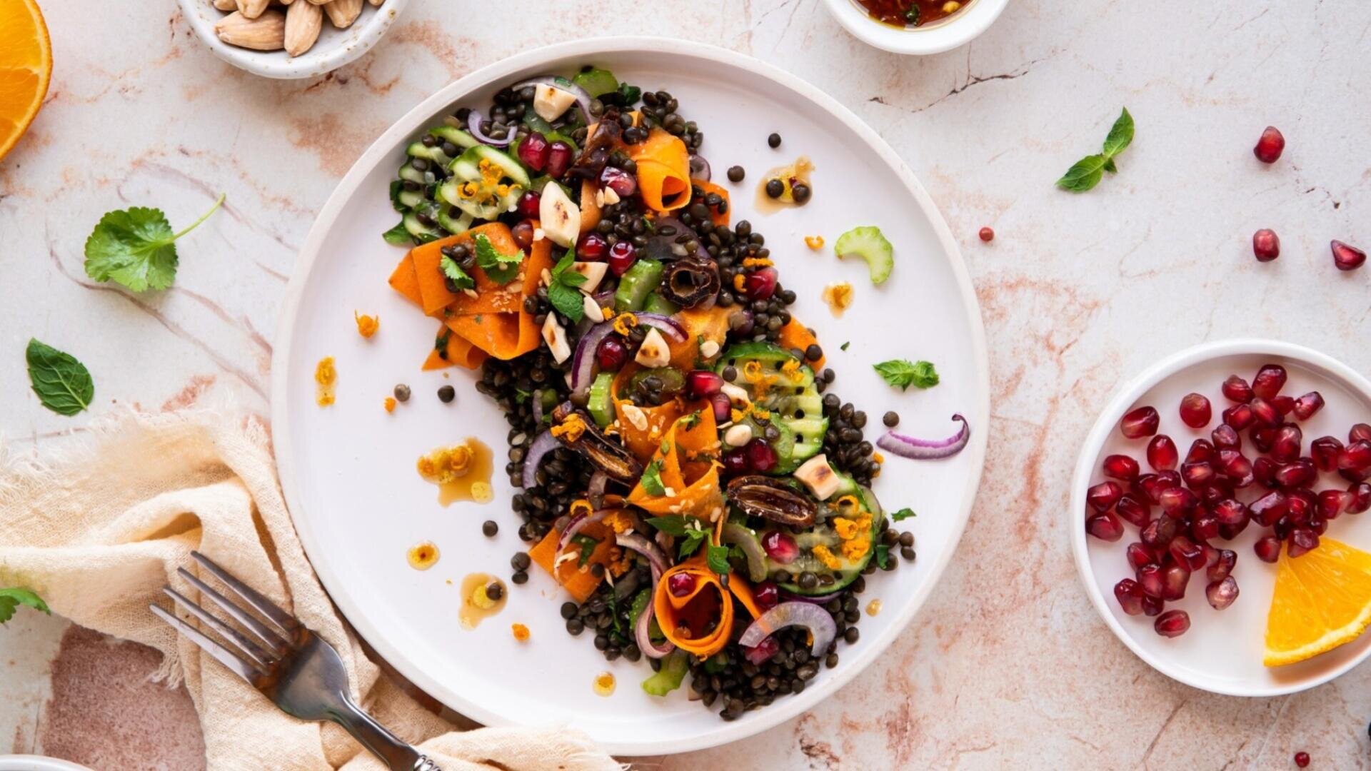 Ein weißer Teller mit einem bunten Linsensalat, garniert mit Karottenscheiben, roten Zwiebeln, Gurke, Granatapfelkernen und Kräutern. Kleine Schälchen mit Nüssen, Granatapfelstücken und Orangenscheiben umgeben den Teller auf einer Marmorplatte.