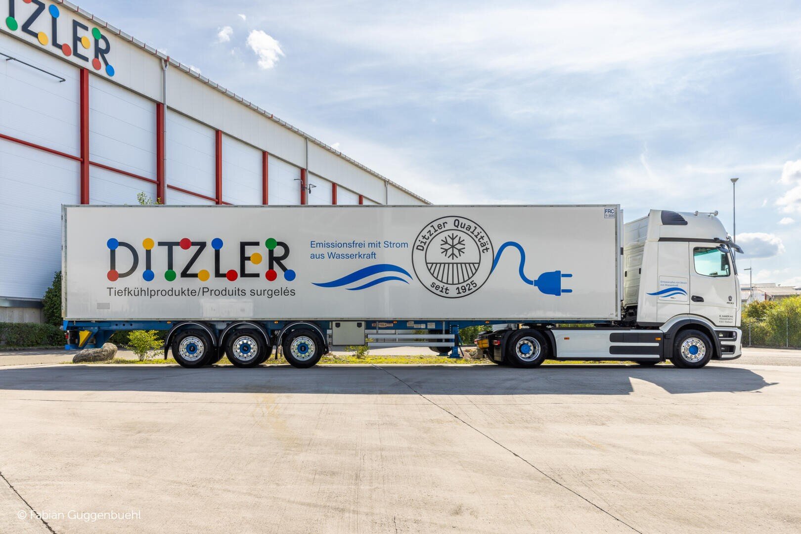 Ein großer weißer Ditzler-LKW mit buntem Logo und blauer Stromsteckergrafik ist vor einer Lagerhalle geparkt. Auf der Seite steht "Emissionsfrei mit Strom aus Wasserkraft", was auf die Verwendung von emissionsfreiem Strom aus Wasserkraft hinweist.