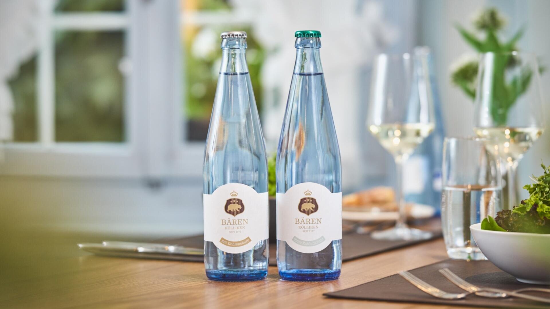 Auf einem gedeckten Esstisch stehen zwei Glasflaschen „Bären“-Sprudelwasser neben Weingläsern, Besteck und einer Salatschüssel, im Hintergrund ist ein helles Fenster zu sehen.