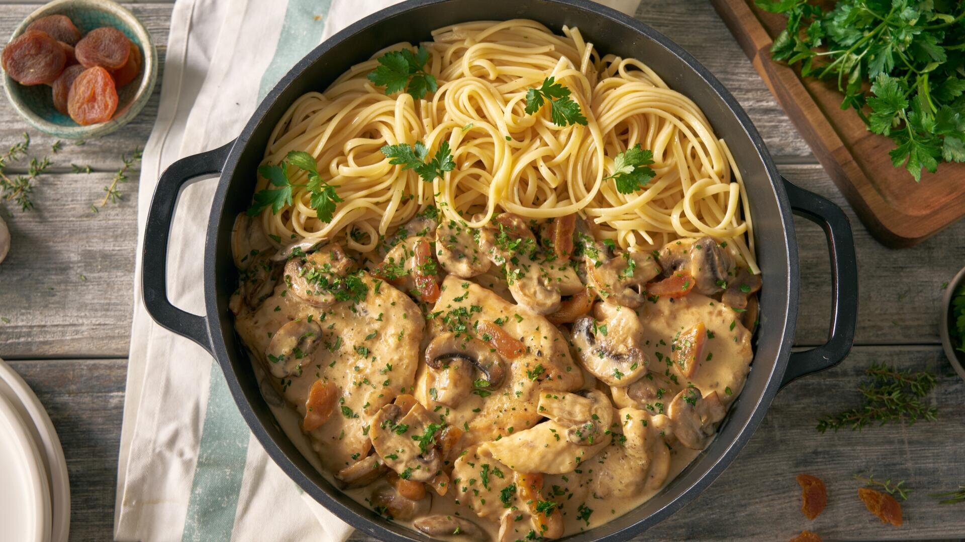 Neben einer Portion einfachen Spaghetti steht eine Pfanne mit cremigem Hähnchenfleisch, Pilzen und Kräutern. Darüber ist gehackte Petersilie gestreut, und auf dem Tisch liegen daneben frische Kräuter sowie ein Holzschneidebrett.