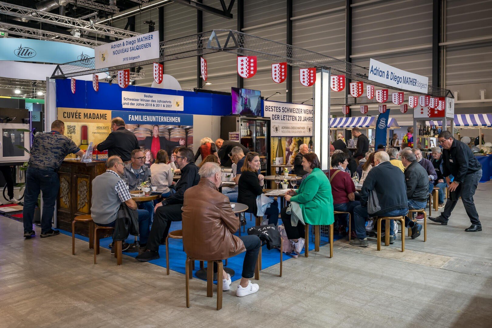 Ein gut besuchter Messestand des Weinguts Adrian & Diego Mathier, an dem Menschen an Tischen sitzen und Wein trinken, während Mitarbeiter die Kunden an einer Theke bedienen.