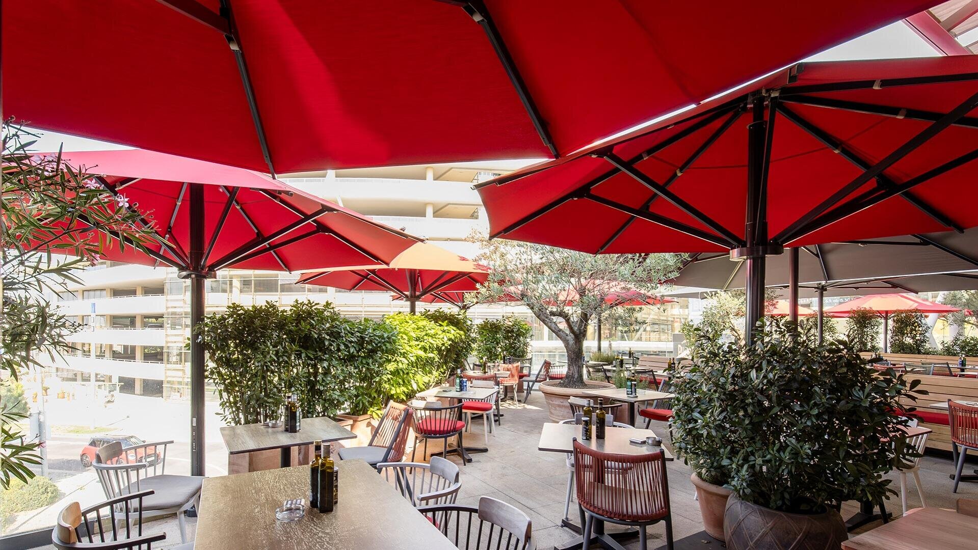 Terrasse eines Restaurants im Freien mit roten Sonnenschirmen, Holztischen und Stühlen, umgeben von grünen Pflanzen. Auf den Tischen stehen Flaschen und Gläser. Im Hintergrund ist ein Parkhaus zu sehen. Helle, sonnige Atmosphäre.