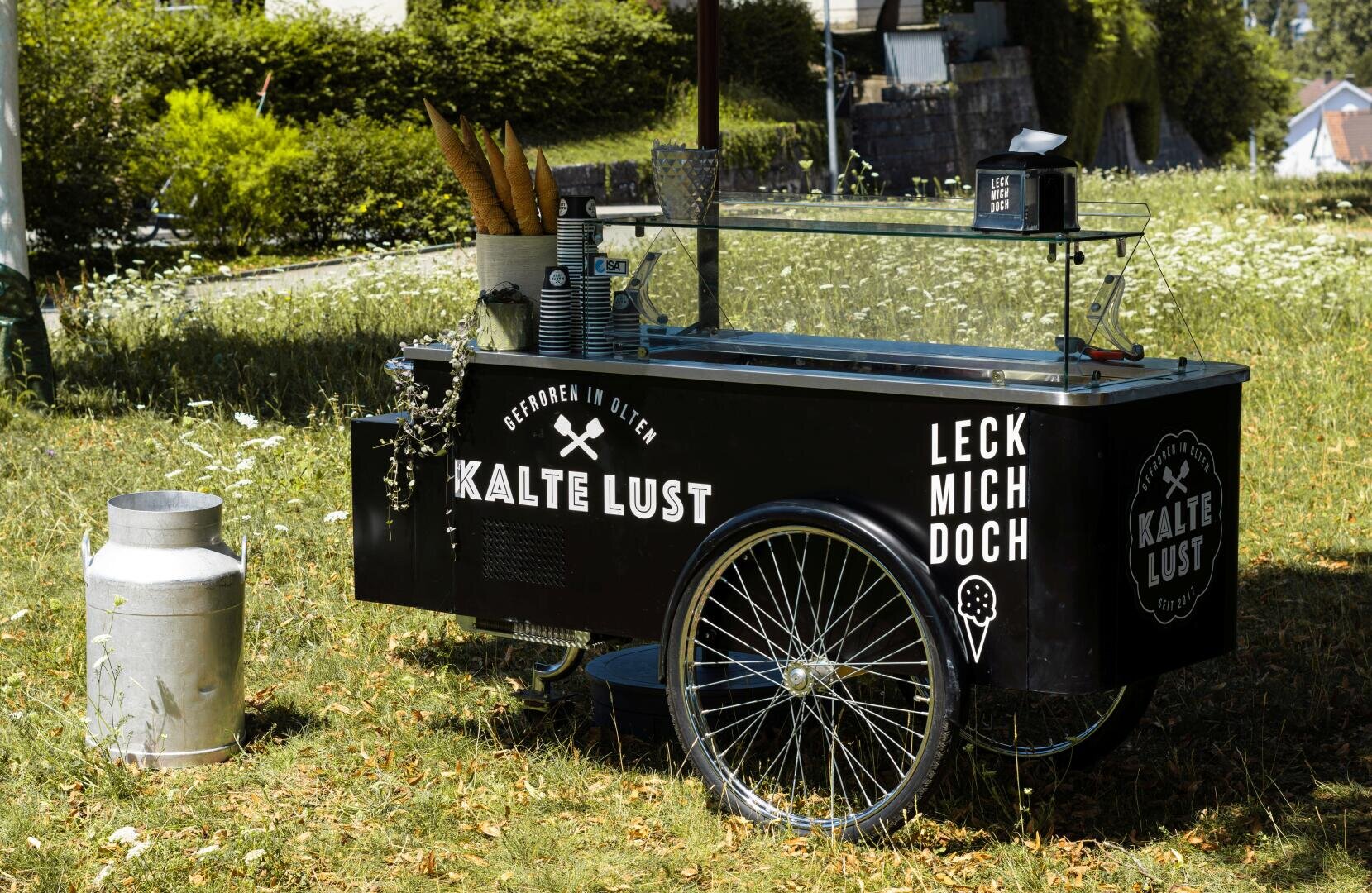 Auf einer Wiese steht ein schwarzer Eiswagen mit der Aufschrift „Kalte Lust“, auf dem Becher, Besteck und Baguettes liegen. Daneben steht eine Milchkanne aus Metall. Im sonnigen Hintergrund sind Bäume und Häuser zu sehen.