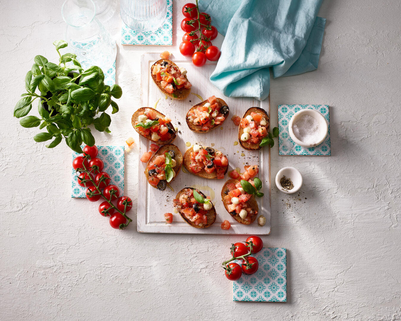 Bruschetta mit Tomatenwürfeln, Basilikum und Käse auf einem weißen Brett, umgeben von Kirschtomaten, Basilikumpflanzen, Salz, Pfeffer, Gläsern und einer blauen Serviette auf einem hellen Untergrund.