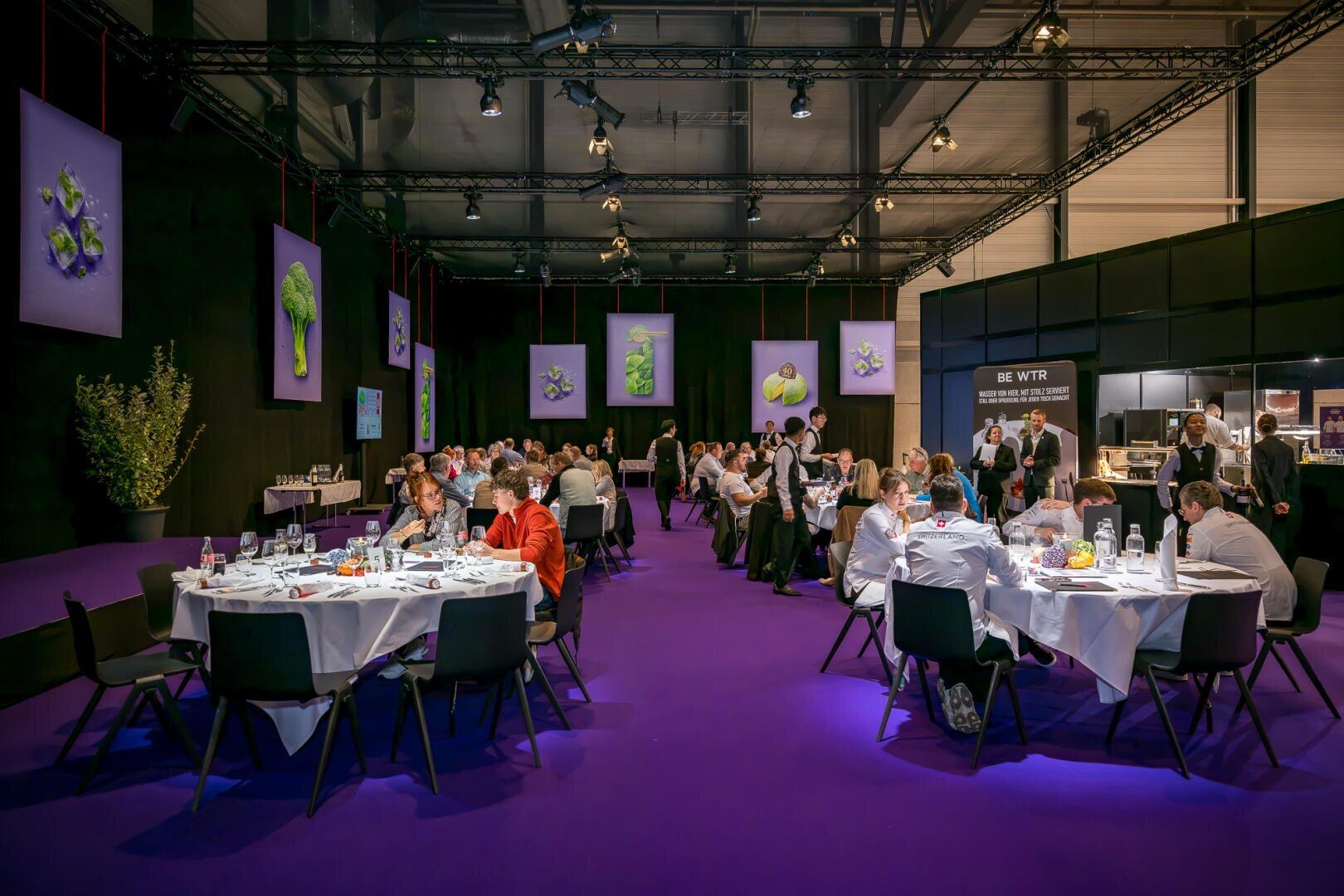 Ein großer Speisesaal mit violettem Boden, in dem sich Menschen an runden Tischen versammelt haben, um an einer Veranstaltung teilzunehmen; an den schwarzen Wänden hängen Plakate mit Gemüsemotiven.