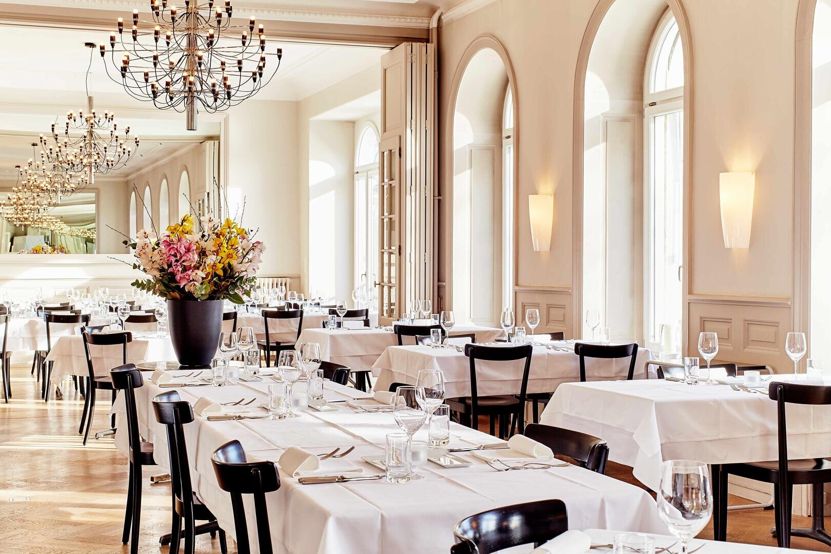 Ein helles, elegantes Restaurant mit weißen Tischdecken, schwarzen Stühlen, Bogenfenstern, Kronleuchtern und einem großen Blumenarrangement in der Mitte. Die Tische sind mit Gläsern und Besteck gedeckt.