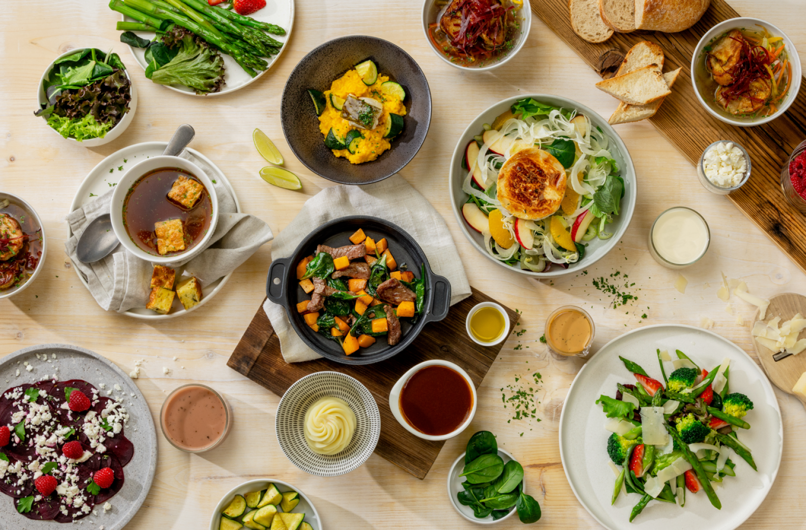 Ein Tisch, reich gedeckt mit verschiedenen Speisen, darunter Salate, Suppen, gebratenes Gemüse, Brot, Soßen und Desserts, die ordentlich auf einer hellen Unterlage angerichtet sind und eine Vielfalt an farbenfrohen, frischen und gesunden Lebensmitteln präsentieren.