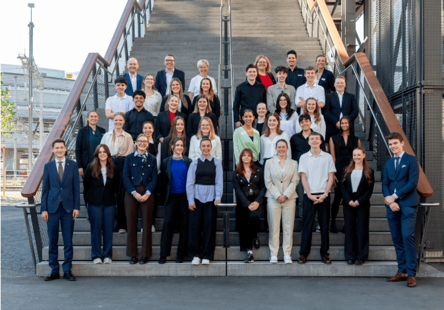 Eine große Gruppe professionell gekleideter Menschen steht an einem sonnigen Tag auf den Stufen einer Freitreppe, lächelt und posiert für ein Gruppenfoto.