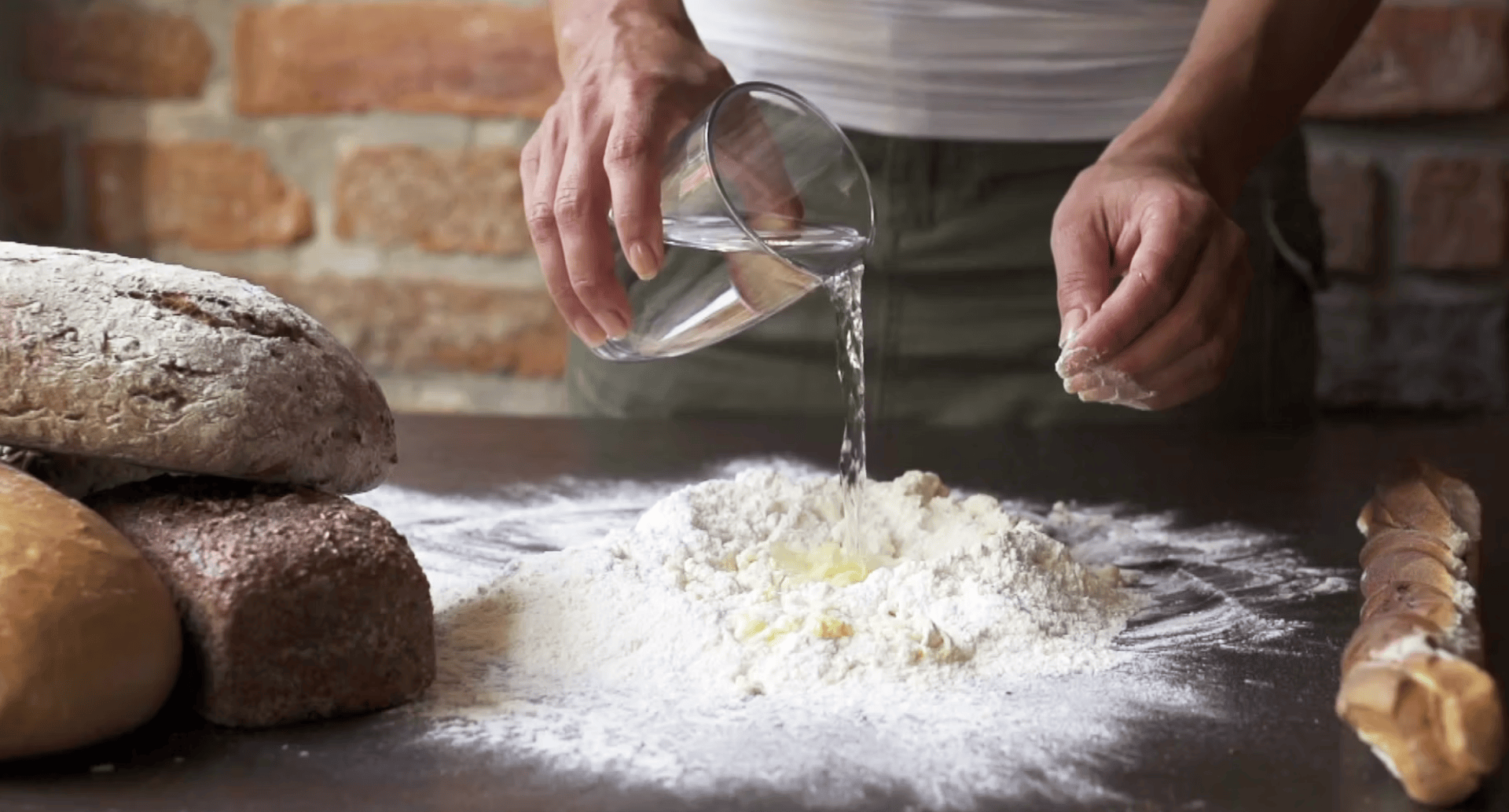 Jemand gießt Wasser aus einem Glas auf einen Mehlhaufen auf einem Tisch, neben dem mehrere Brotlaibe liegen.