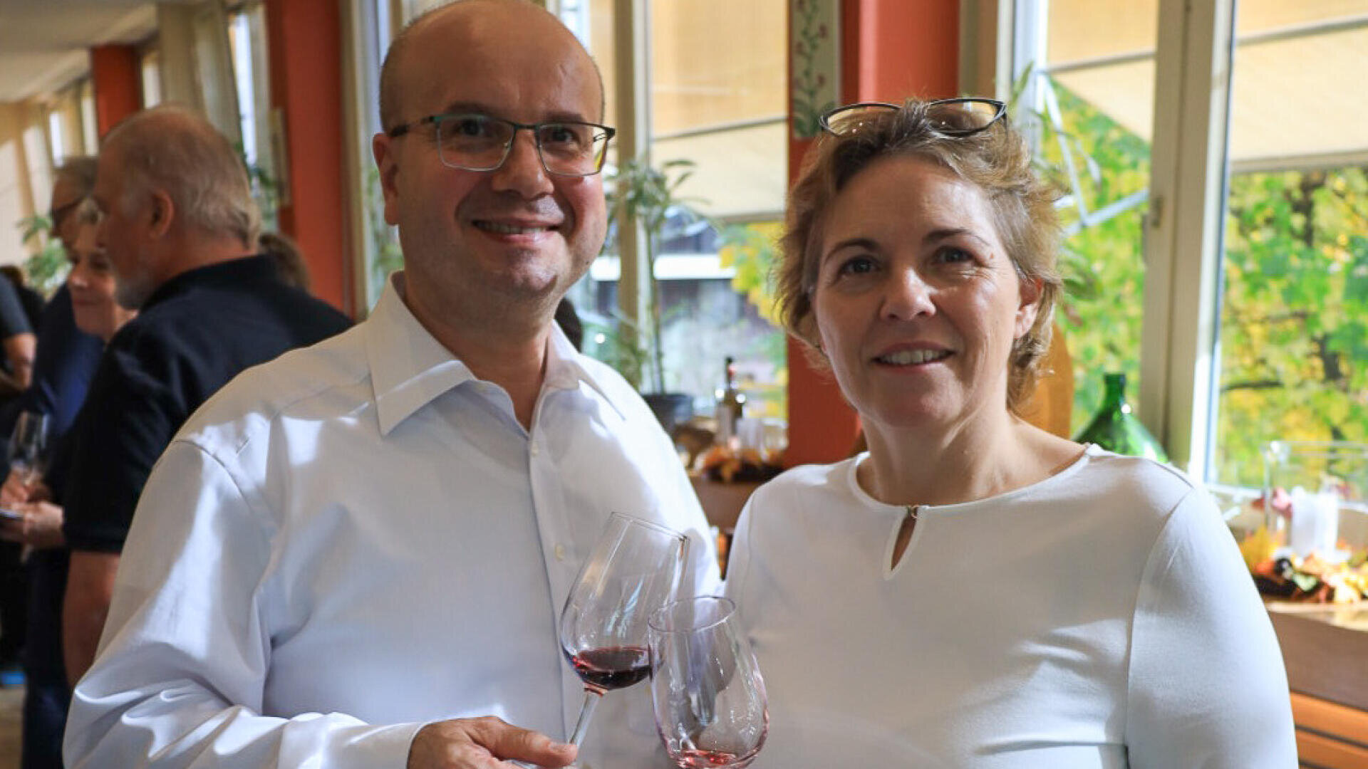 Ein Mann und eine Frau, beide in weißen Oberteilen, lächeln und halten Weingläser mit Rotwein in einem sonnendurchfluteten Raum in der Hand. Im Hintergrund sind weitere Personen und ein Tisch mit Essen zu sehen.