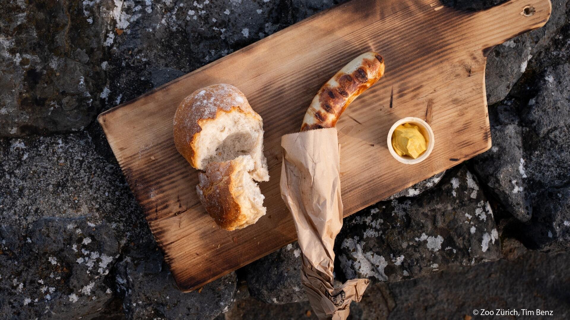 Eine teilweise in Papier eingewickelte Grillwurst, ein knuspriges Brötchen und ein kleines Schälchen Senf liegen auf einem Holzbrett, das auf rauen grauen Steinen steht.