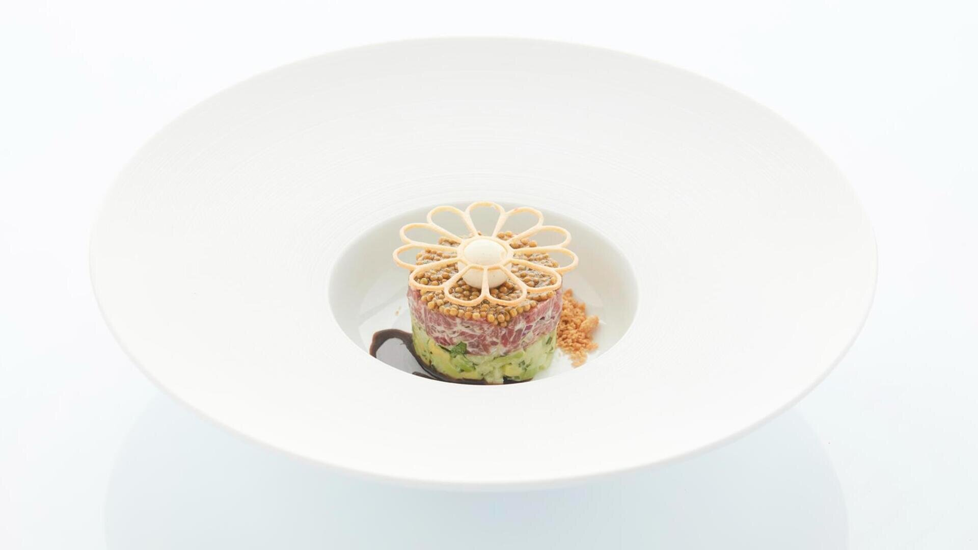 Ein Gourmetgericht aus geschichteten Gemüse- und Fleischschichten, gekrönt von einer dekorativen, blumenförmigen Kruste, serviert in der Mitte einer breiten weißen Schüssel, flankiert von einer Prise Bröseln.