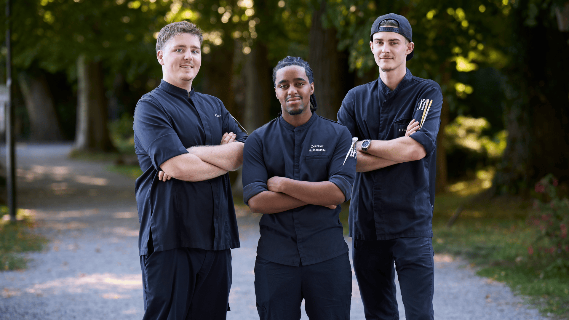 Drei Köche in schwarzen Uniformen stehen draußen und haben die Arme verschränkt.