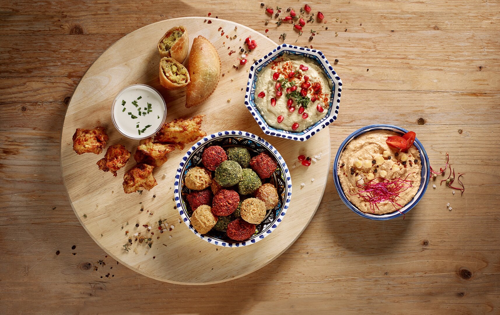 Eine Holzplatte mit bunten Falafeln, zwei Schalen Hummus mit verschiedenen Beilagen, Frühlingsrollen, Hähnchenspieße und einer kleinen Schale mit Dip, angerichtet auf einem rustikalen Holztisch.