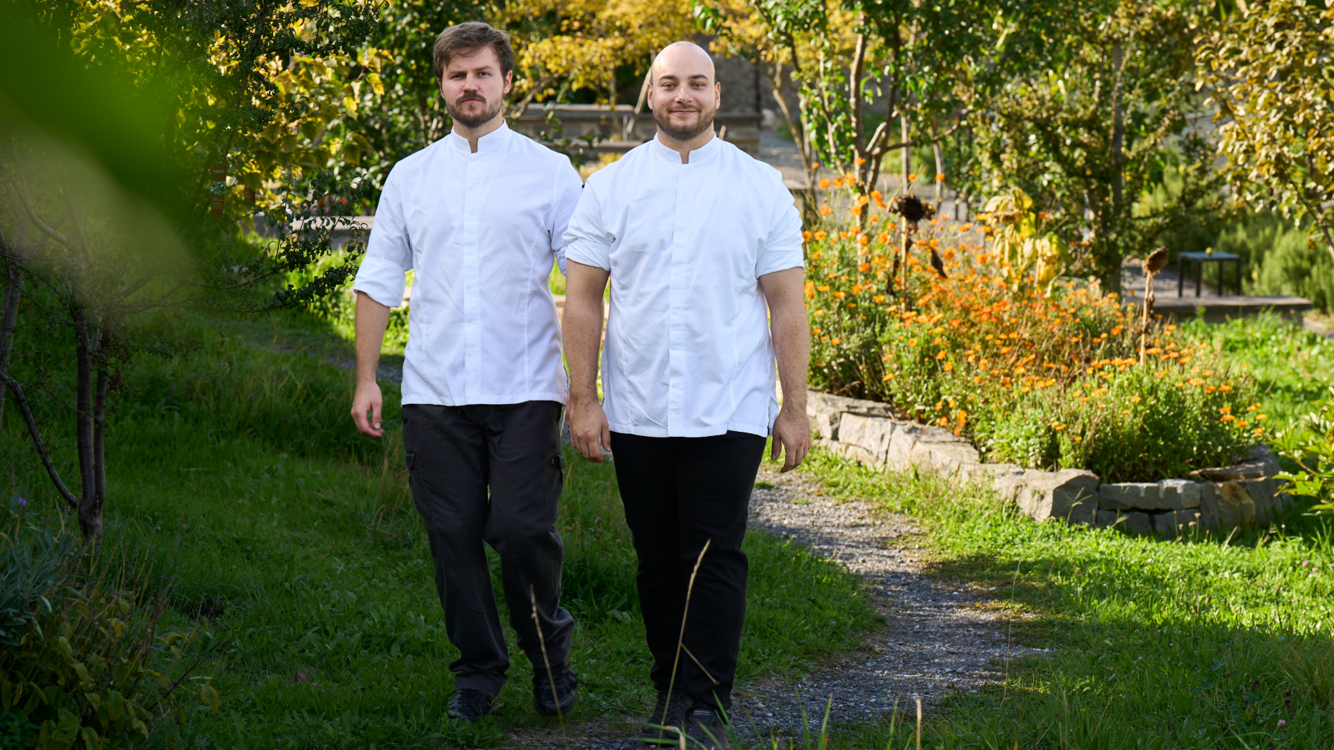 Zwei Köche in weißen Uniformen, die nebeneinander einen Gartenweg entlanggehen.