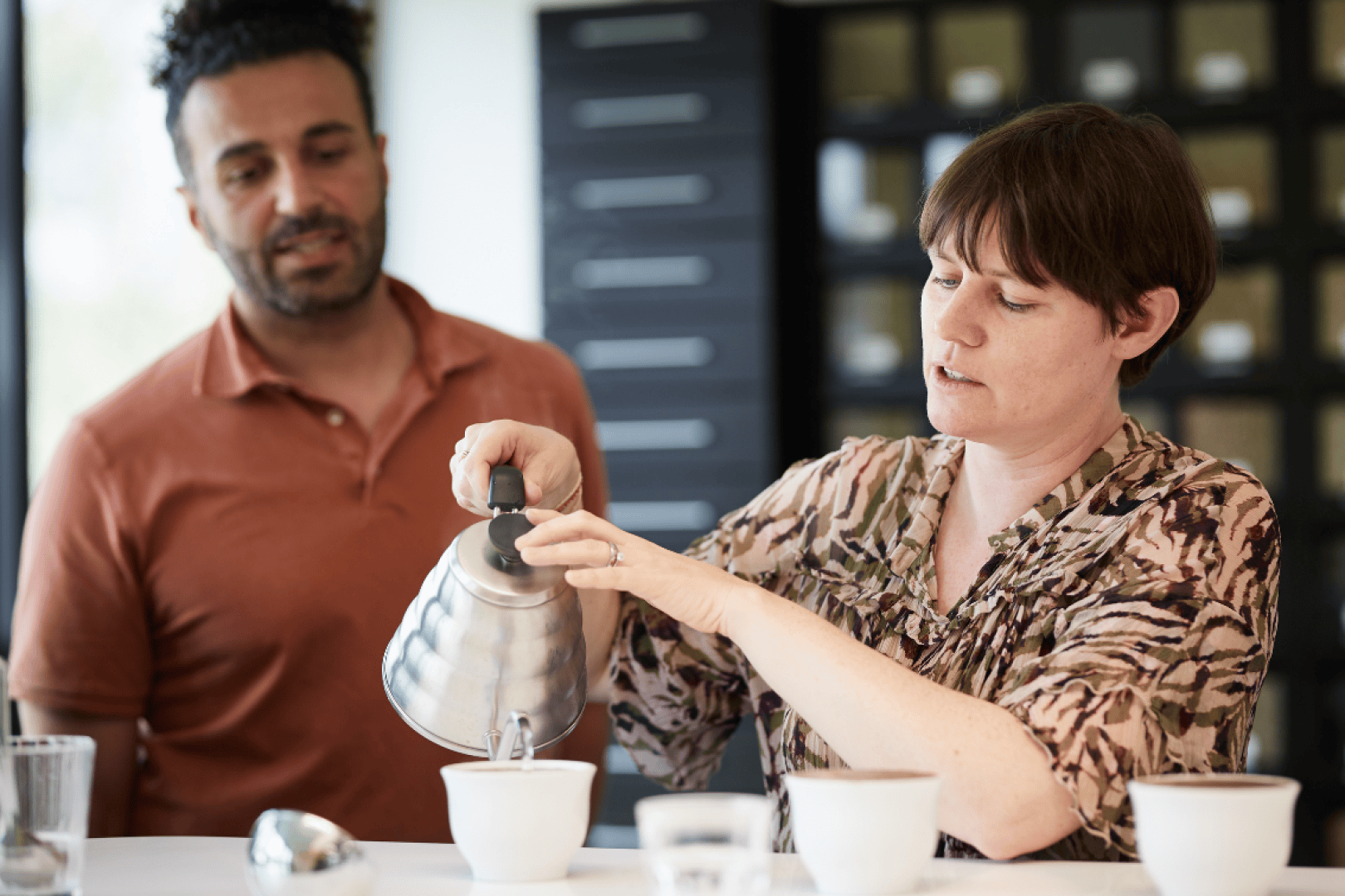 Eine Frau gießt heißes Wasser aus einem Metallkessel in eine weiße Tasse, während ein Mann neben ihr steht und aufmerksam zusieht. Beide befinden sich in einem Raum, und im Hintergrund sind Regale mit Gläsern zu sehen.