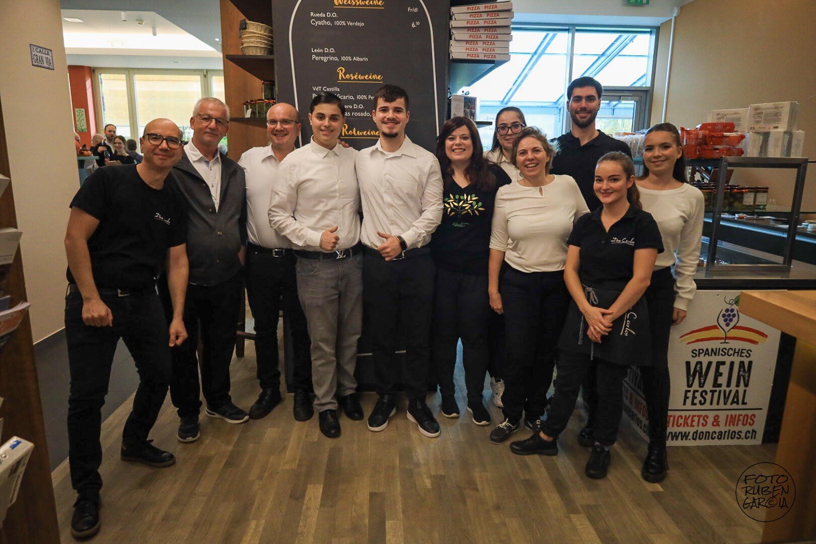 Eine Gruppe von zwölf lächelnden Menschen, von denen einige schwarze und andere weiße Hemden tragen, steht gemeinsam in einem Restaurant und posiert für ein Gruppenfoto in der Nähe einer Theke und einer aufgehängten Speisekarte.