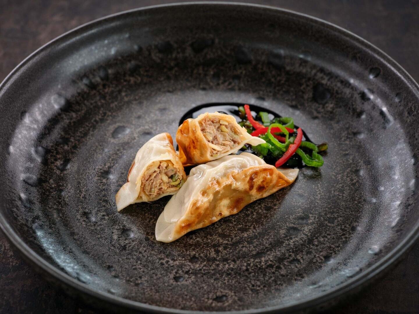Zwei in der Pfanne gebratene Teigtaschen, von denen eine halbiert ist, serviert mit einer dunklen Dip-Sauce, Frühlingszwiebeln und Chilischeiben auf einem dunklen Teller.