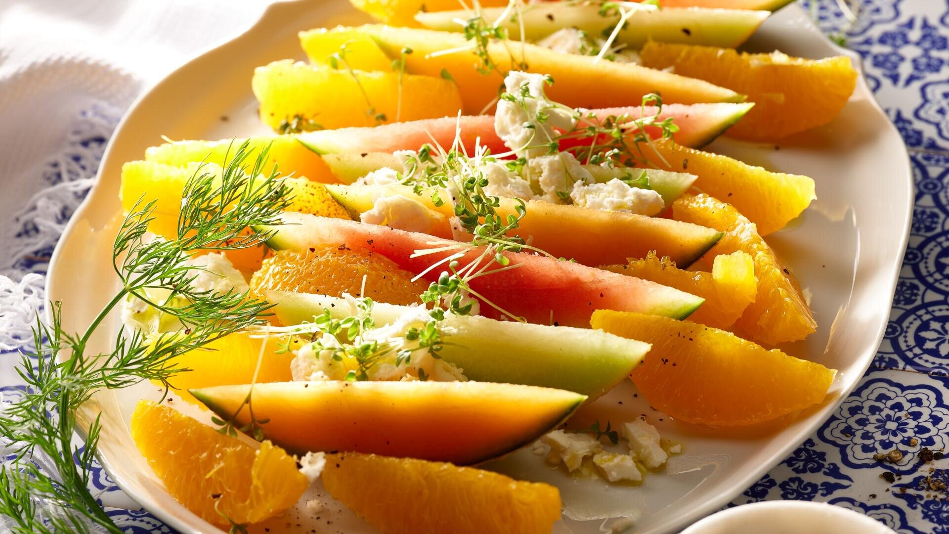 Auf einem Teller, der auf einer blau-weiß gemusterten Tischdecke liegt, sind in Scheiben geschnittene Wassermelone, Cantaloupe-Melone und Orangenschnitze angerichtet, garniert mit zerbröckeltem Käse, frischen Kräutern und Microgreens.