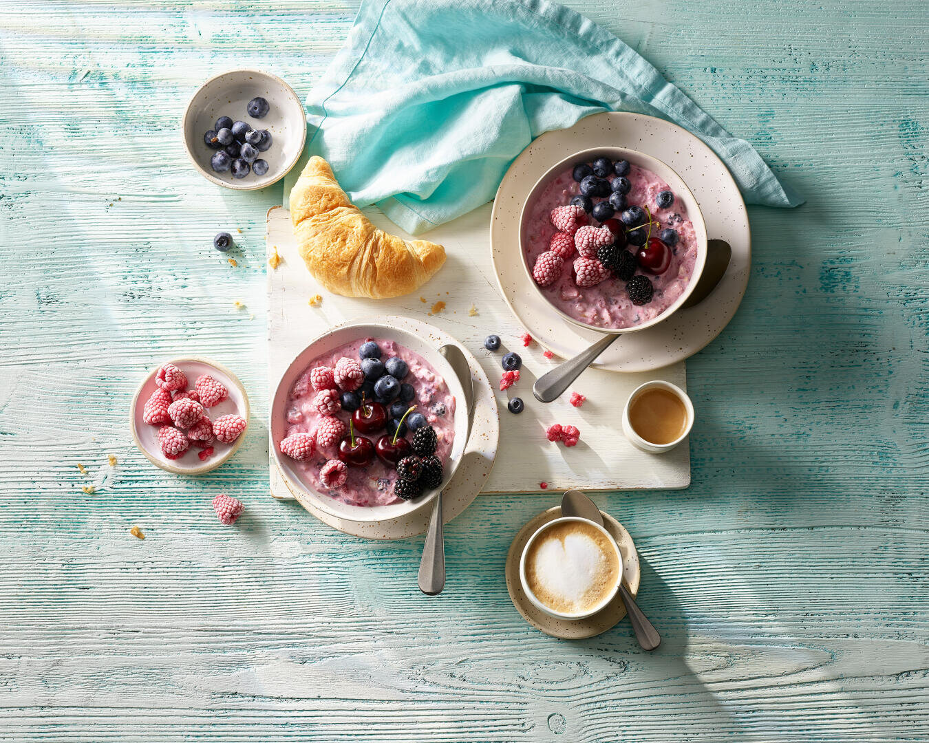 Zwei Schalen Beerenjoghurt mit verschiedenen Beeren, ein Croissant, eine Tasse Kaffee, eine kleine Schale mit Heidelbeeren und eine Serviette auf einem hellblauen Holztisch.