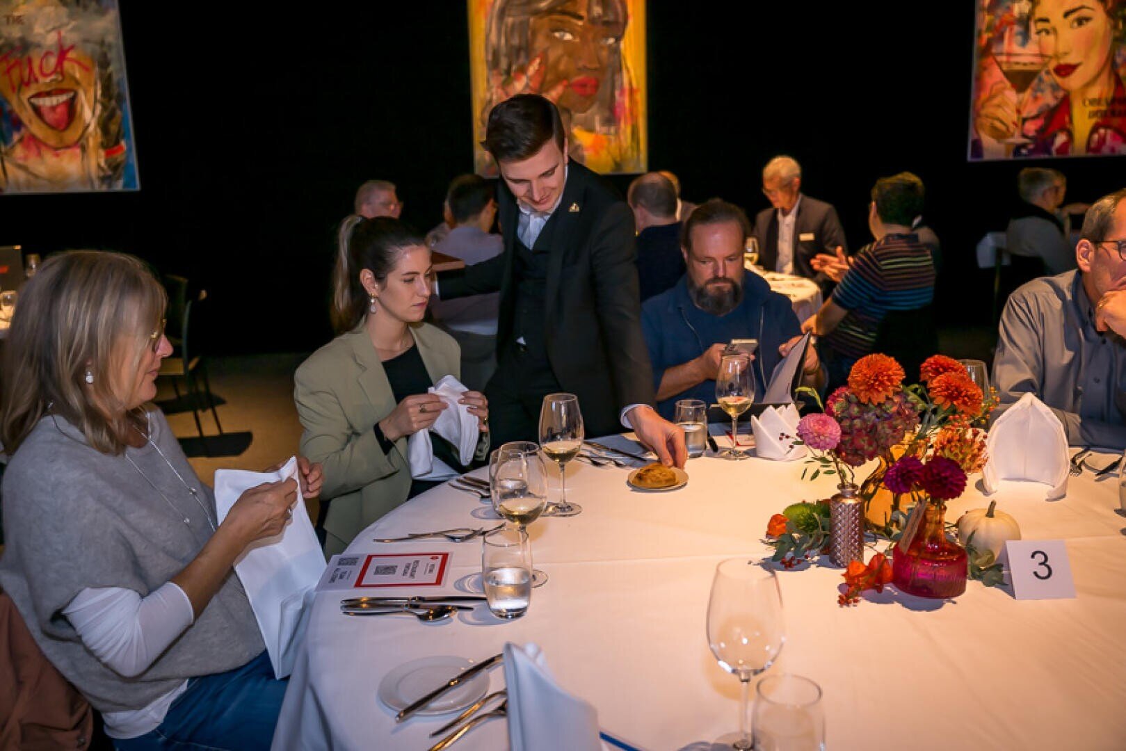 Ein Kellner serviert den Gästen, die an einem runden Tisch mit Blumengestecken und festlichem Geschirr sitzen, bei einem festlichen Abendessen Brot. Im Hintergrund sind Kunstwerke mit Gesichtern zu sehen.