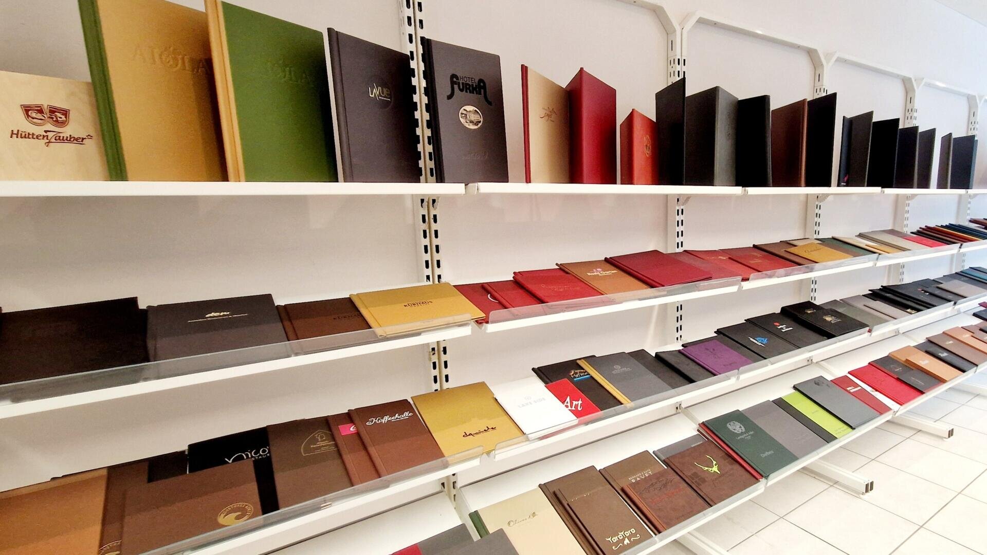 Auf drei weißen Regalen sind verschiedene geschlossene Bücher und Speisekarten in unterschiedlichen Farben und Größen ausgestellt, die ordentlich in einem hell beleuchteten Raum mit weißem Boden und weißen Wänden angeordnet sind.