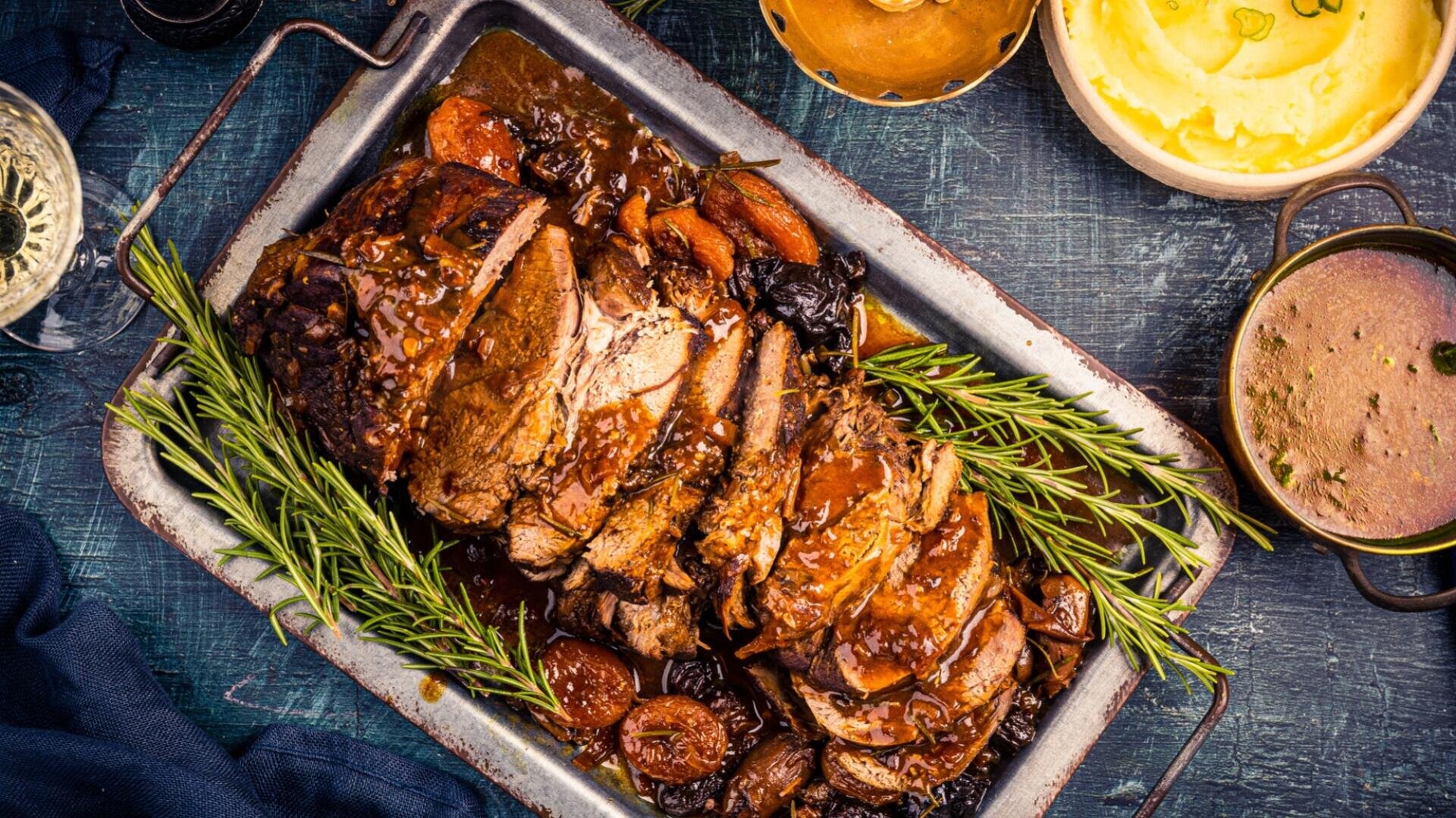 In Scheiben geschnittenes Roastbeef mit Bratensoße, Möhren, Pflaumen und frischem Rosmarin auf einem Metalltablett, daneben Kartoffelpüree und Soße, auf einer rustikalen, dunklen Unterlage.