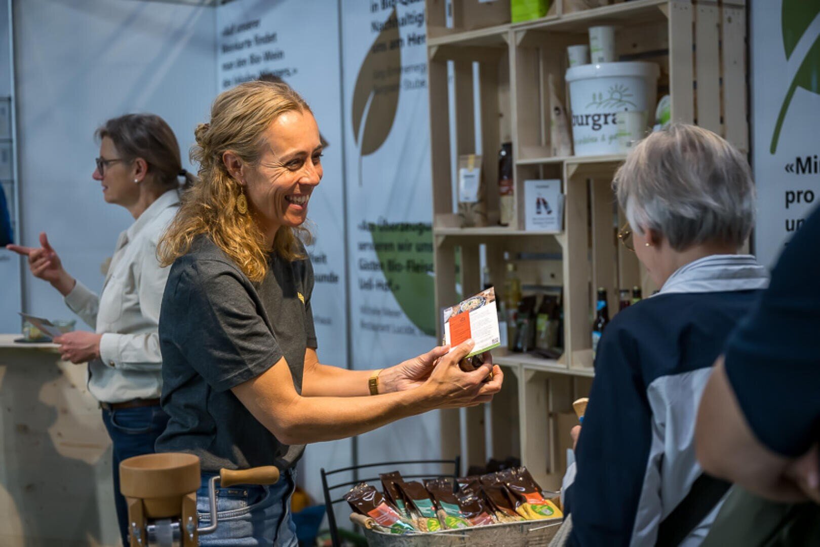 Eine lächelnde Frau zeigt einem Kunden an einem Stand mit Warenregalen ein Produkt. Im Hintergrund ist eine weitere Person zu sehen, die mit jemandem außerhalb des Bildausschnitts spricht.