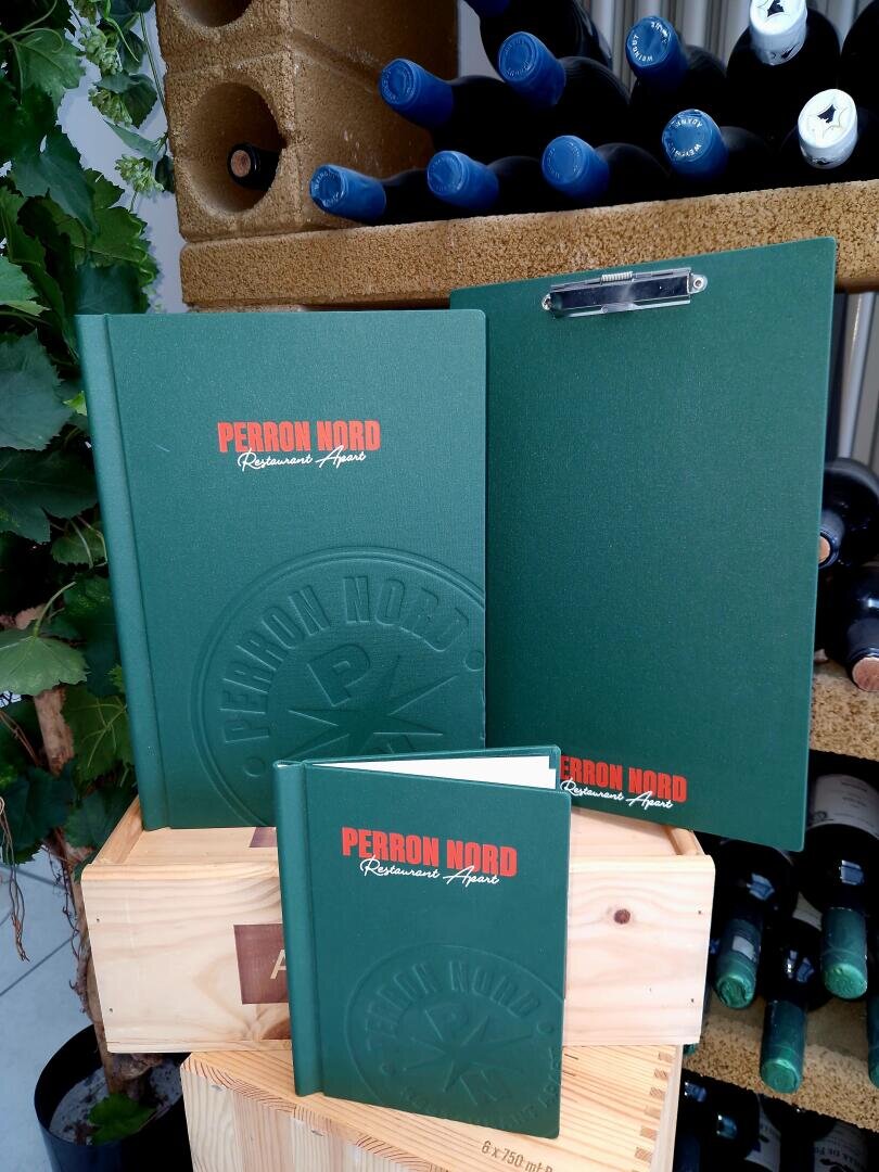 Auf Holzkisten sind drei grüne Speisekarten des „Perron Nord Ristorante Aperto“ sowie ein Klemmbrett zu sehen, im Hintergrund sind Weinflaschen und Weinreben zu erkennen.
