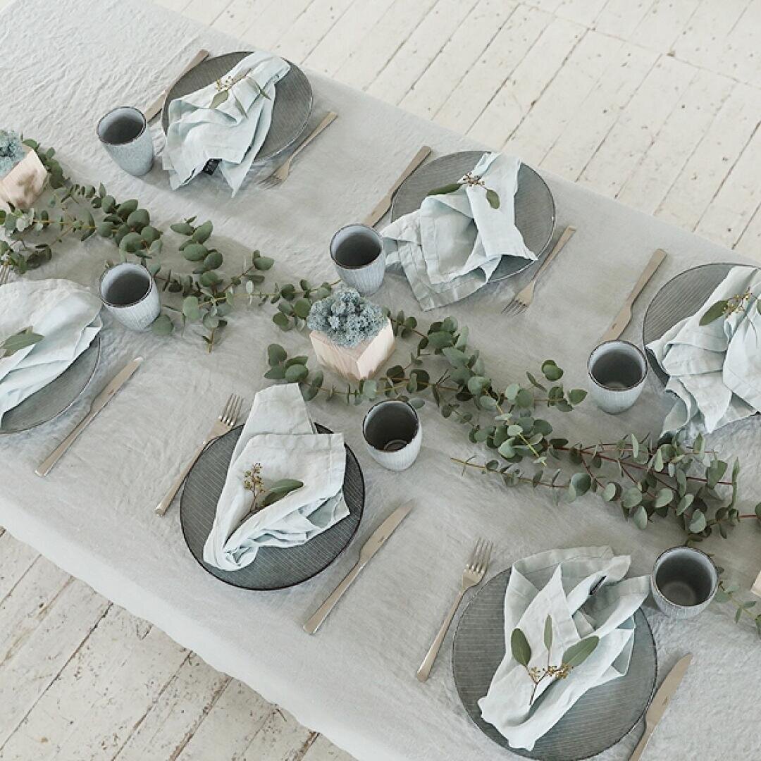 Elegante Tischdekoration mit grauen Tellern, silbernem Besteck, hellen, mit Grün verzierten Servietten, grauen Tassen und einer Eukalyptusgirlande als Tischschmuck auf einer hellen Tischdecke.