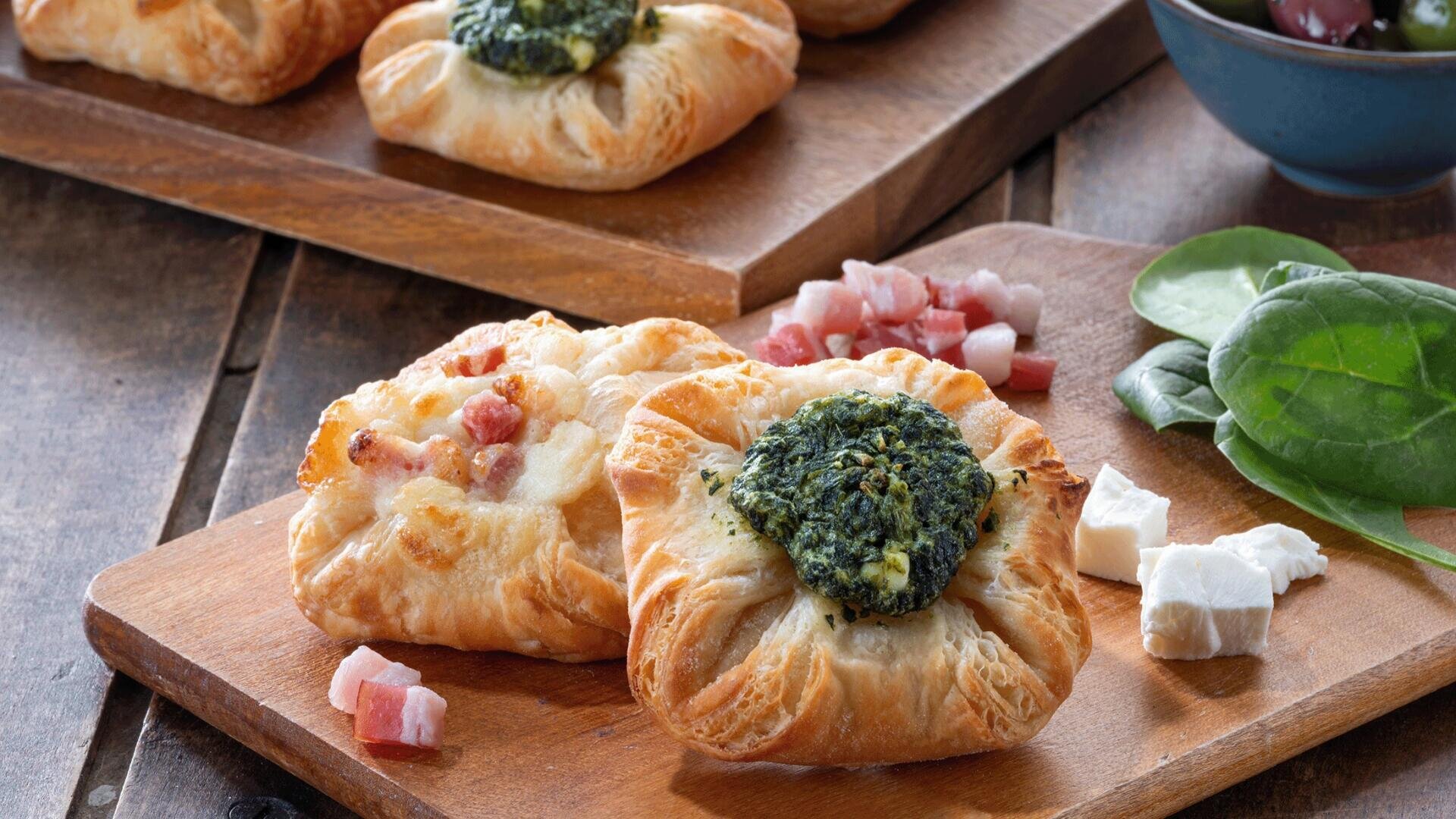 Zwei Blätterteigtörtchen auf einem Holzbrett, eines mit Spinat und Käse belegt, das andere mit Schinkenwürfeln. Daneben liegen Spinatblätter, Käse und Schinkenstücke.