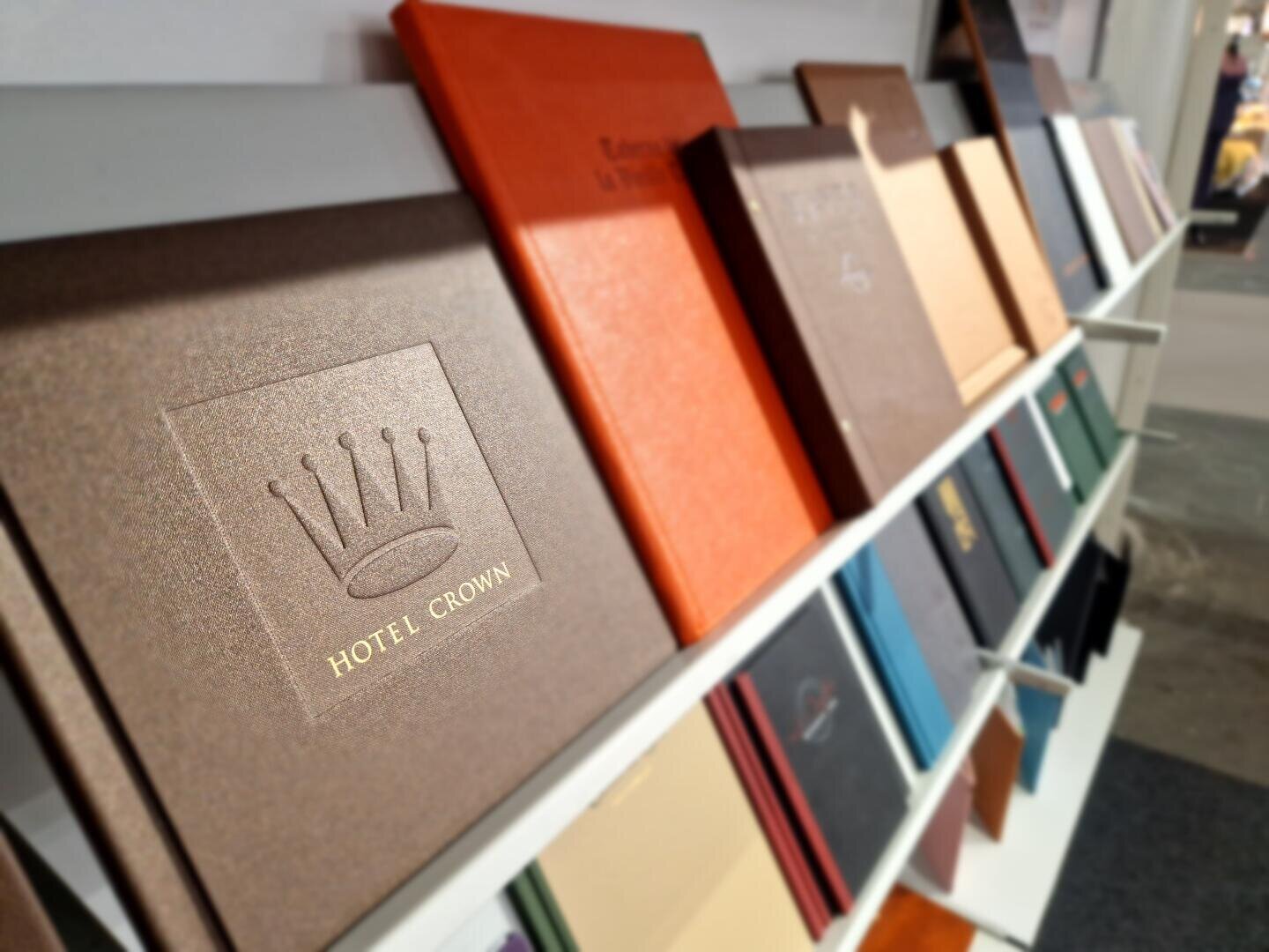 Eine Nahaufnahme von Büchern in einem Regal, wobei der Fokus auf einem braunen Buch liegt, auf dem eine Krone und die Aufschrift „HOTEL CROWN“ eingeprägt sind. Andere Bücher in verschiedenen Farben sind im Hintergrund unscharf zu sehen.