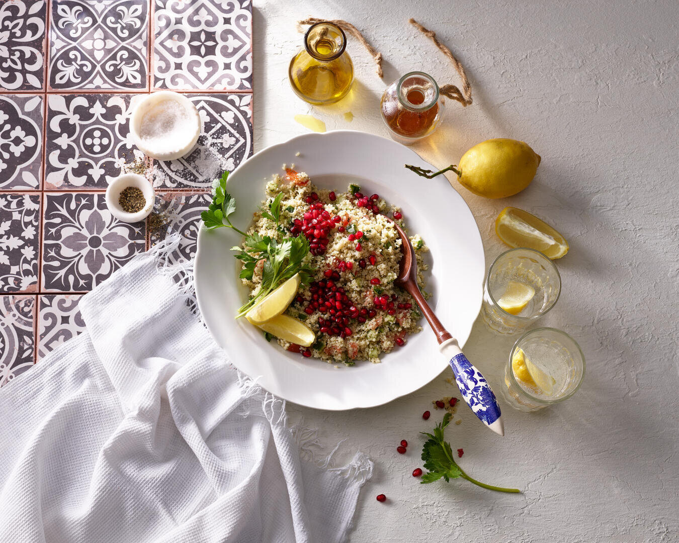 Eine Schüssel Quinoa-Salat mit Granatapfel, Petersilie und Zitronenschnitzen steht auf einer weißen Tischdecke neben Getränken, Gewürzen, Olivenöl, Honig und einer ganzen Zitrone auf einer gefliesten, strukturierten Oberfläche.