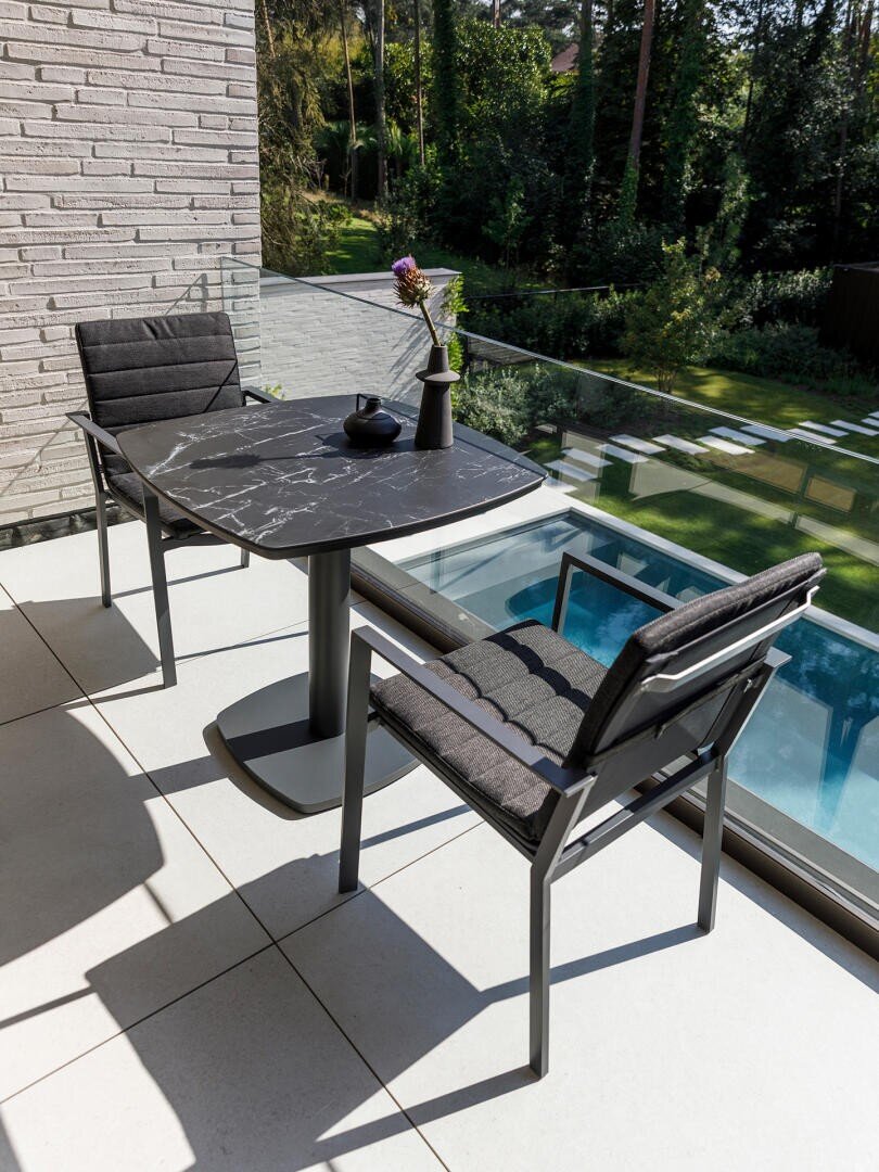 Ein moderner Balkon mit zwei schwarzen, gepolsterten Stühlen und einem Tisch aus schwarzem Marmor. Auf dem Tisch stehen eine einzelne Vase mit einer Blume, eine kleine Schale und eine Tasse. Durch das Glasgeländer blickt man auf einen grünen Hinterhof.