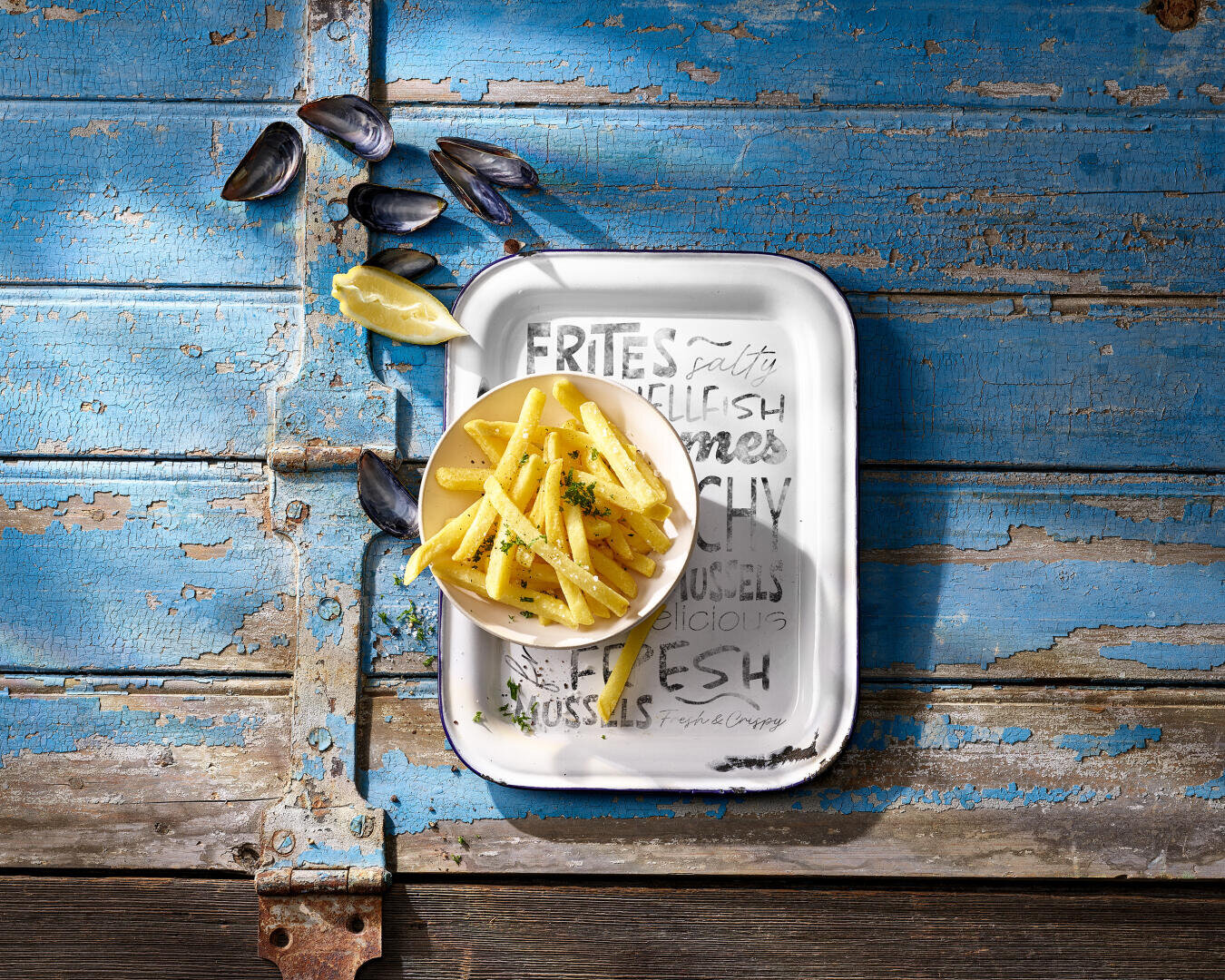 Auf einer abgenutzten blauen Holzplatte steht ein Teller mit Pommes frites mit Kräutern, garniert mit Zitronenschnitzen und umgeben von leeren Muschelschalen.