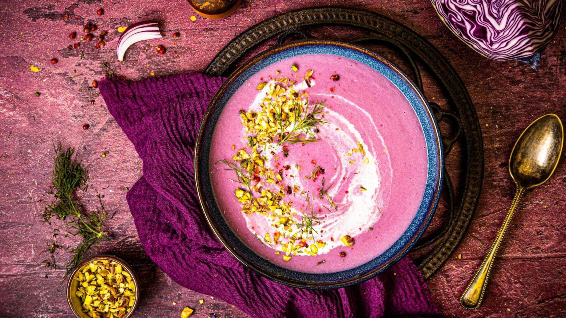Eine Schüssel mit cremiger, violetter Suppe, garniert mit Kräutern und gehackten Nüssen, auf einem violetten Tuch serviert, daneben ein Löffel und weitere Zutaten – das Ganze auf einer strukturierten, rötlichen Oberfläche.