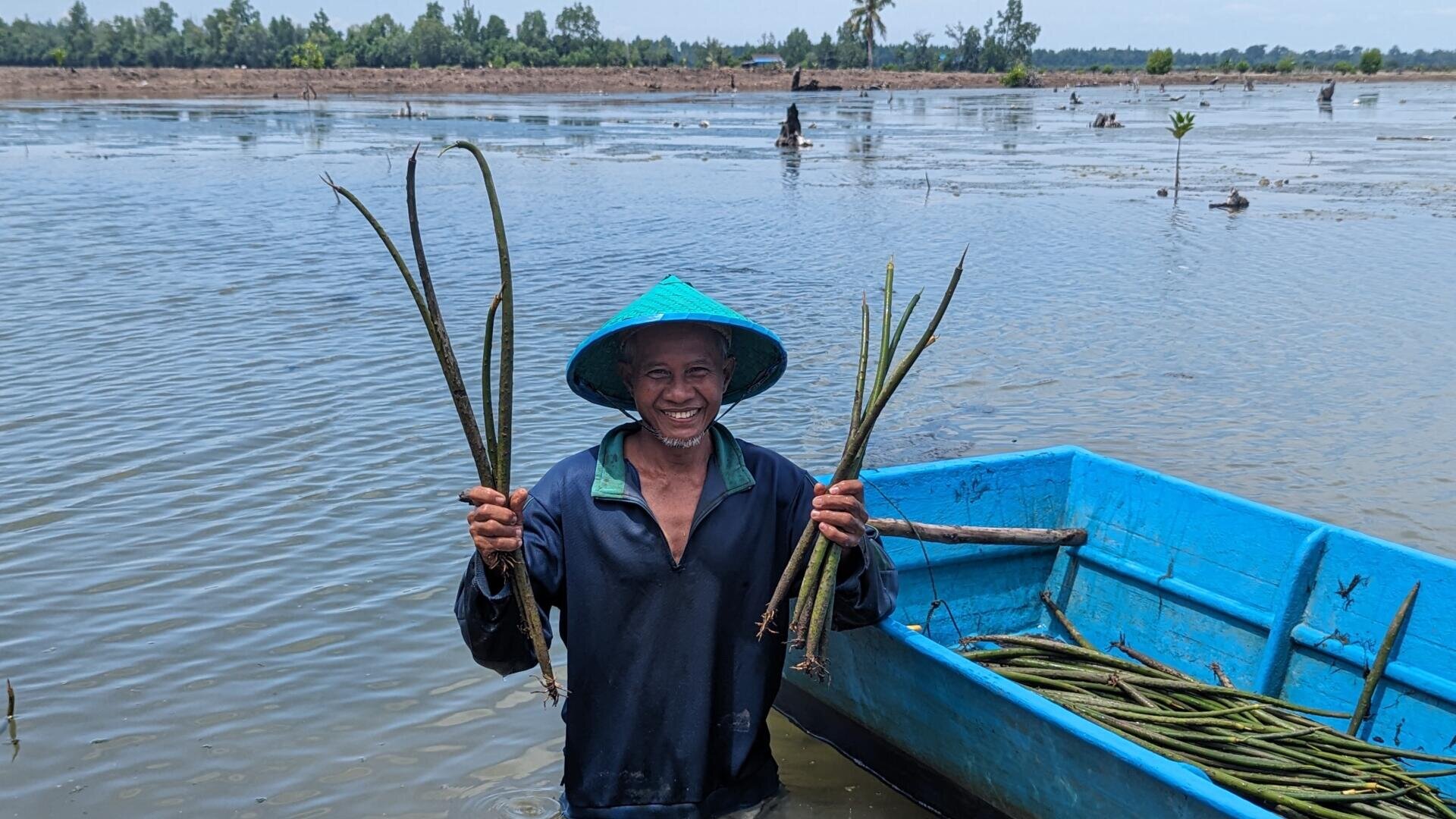 Eine lächelnde Person mit blauem Hut und blauem Hemd steht im seichten Wasser und hält neben einem blauen Boot in beiden Händen lange grüne Pflanzen in den Händen. Im Hintergrund sind Bäume und Vegetation zu sehen.