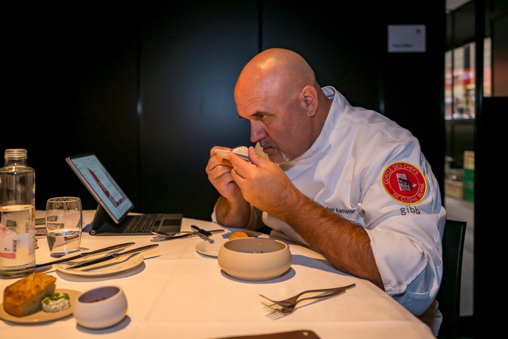 Ein Koch in weißer Kochkleidung begutachtet an einem Restauranttisch aufmerksam das Essen, um ihn herum stehen ein Laptop, Teller, Gläser und Brot.