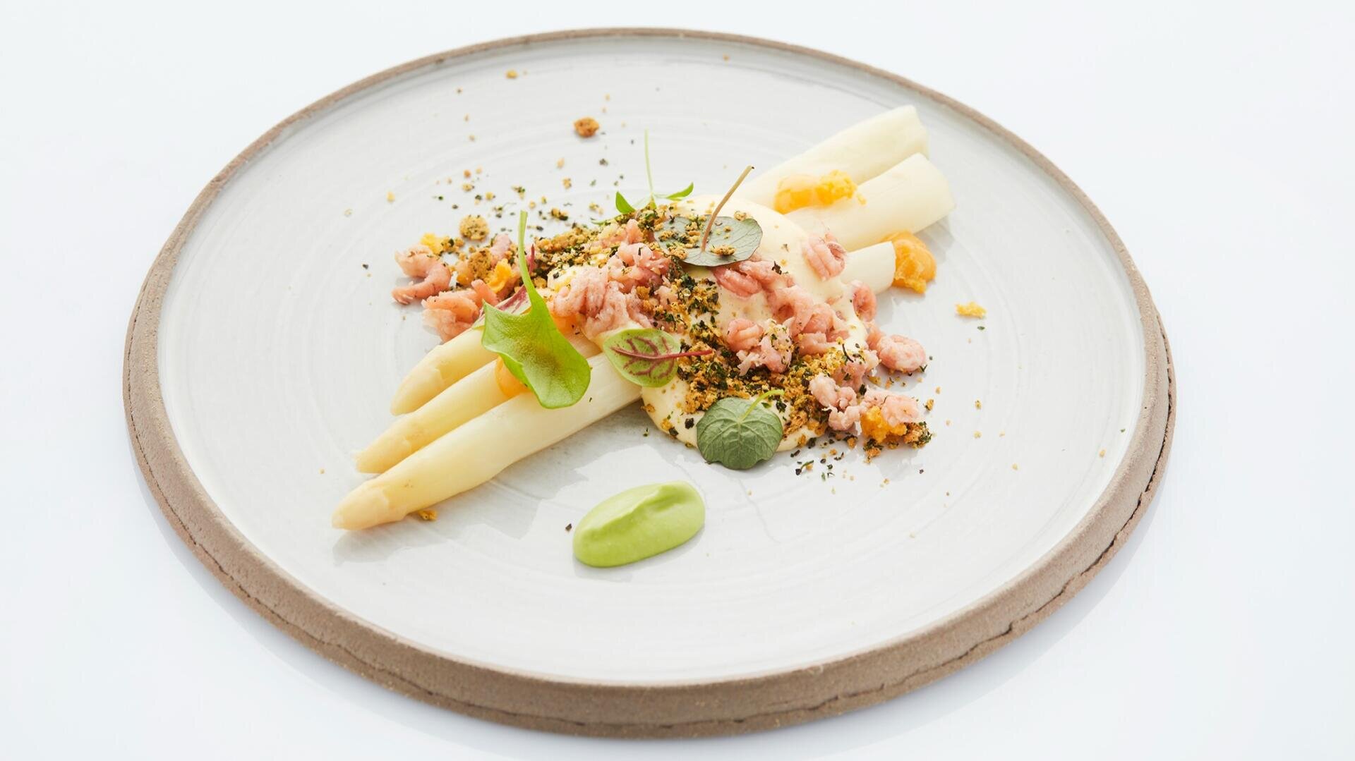 Drei weiße Spargelstangen, garniert mit fein gehackten Meeresfrüchten, Microgreens, Streuseln und einem Klecks grüner Soße, serviert auf einem runden weißen Teller.