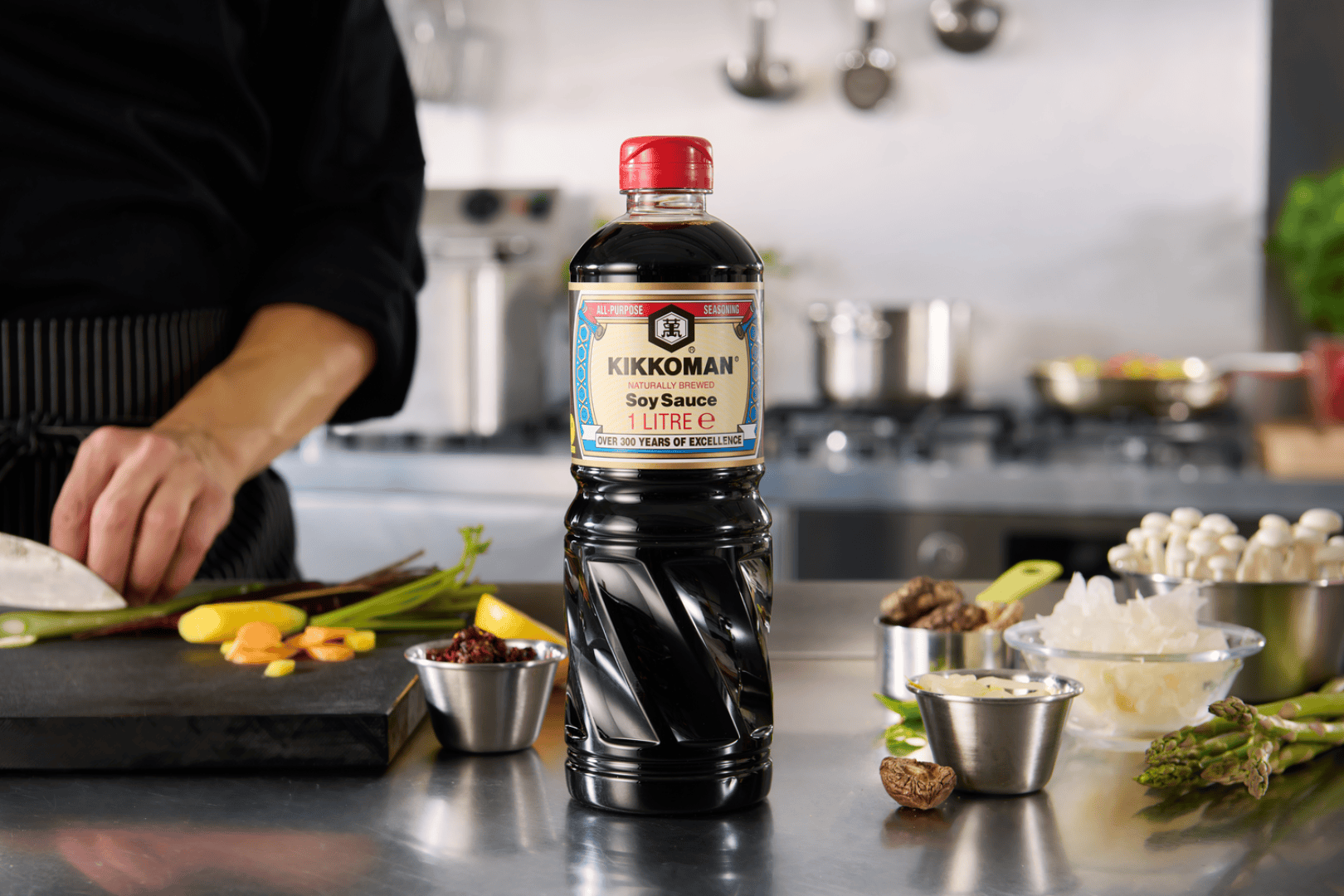 Eine Flasche Kikkoman-Sojasauce steht auf einer Küchenarbeitsplatte neben geschnittenem Gemüse, Pilzen und anderen Zutaten, während im Hintergrund eine Person Gemüse schneidet.