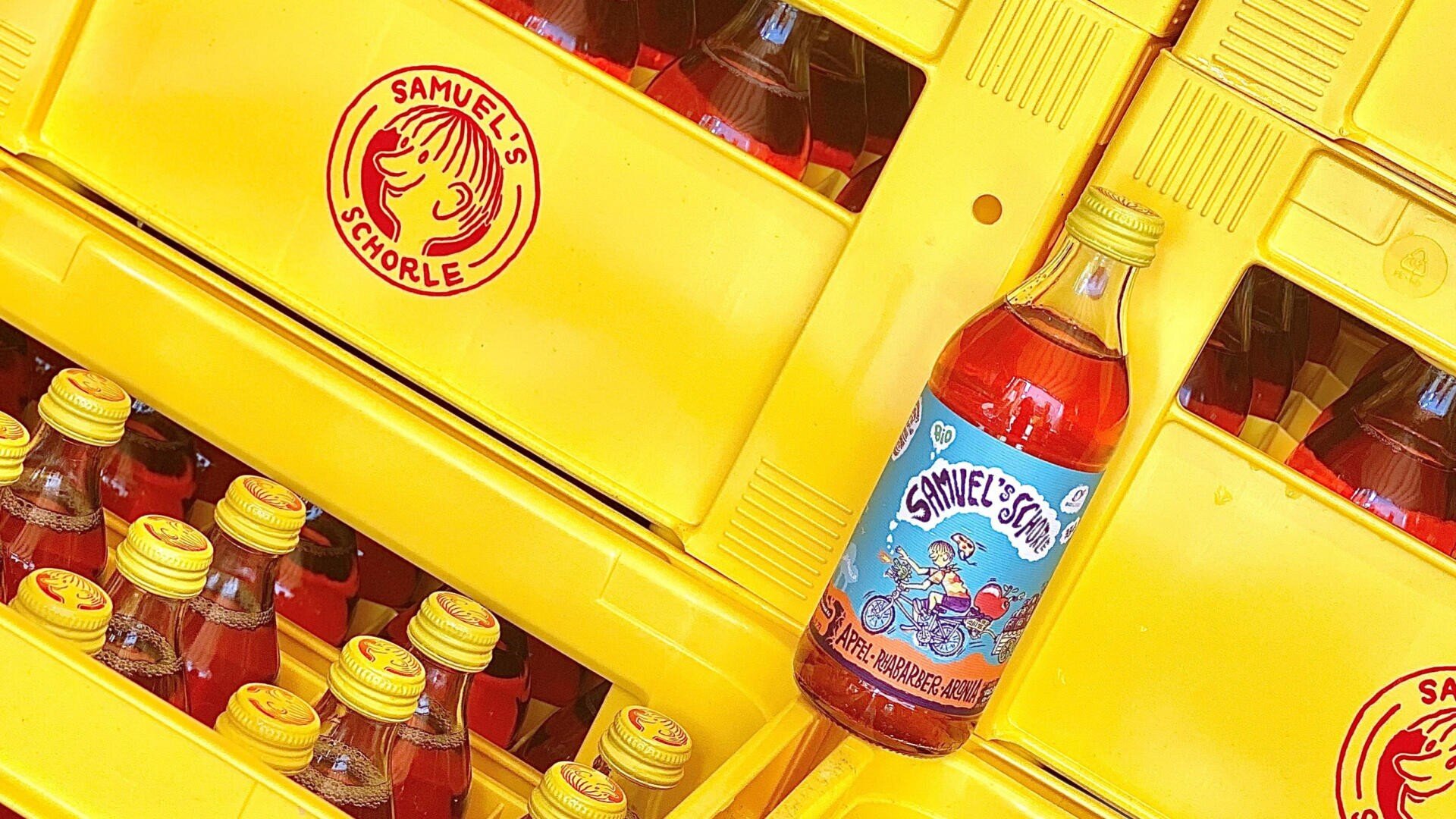 Flaschen „Samuels Schorle“ mit roter Flüssigkeit sind in leuchtend gelben Kisten gestapelt. Eine Flasche steht ganz vorne und zeigt ein buntes Etikett mit Comicfiguren, die Fahrrad fahren.