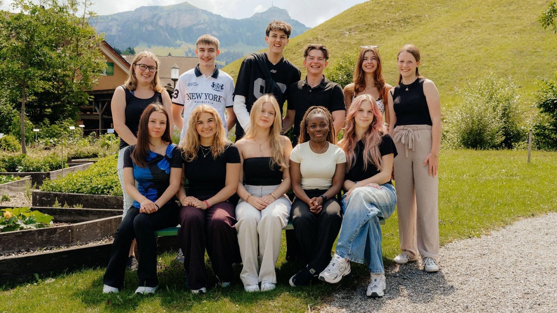 Eine Gruppe von elf jungen Erwachsenen posiert im Freien vor einem grasbewachsenen Hügel für ein Foto.