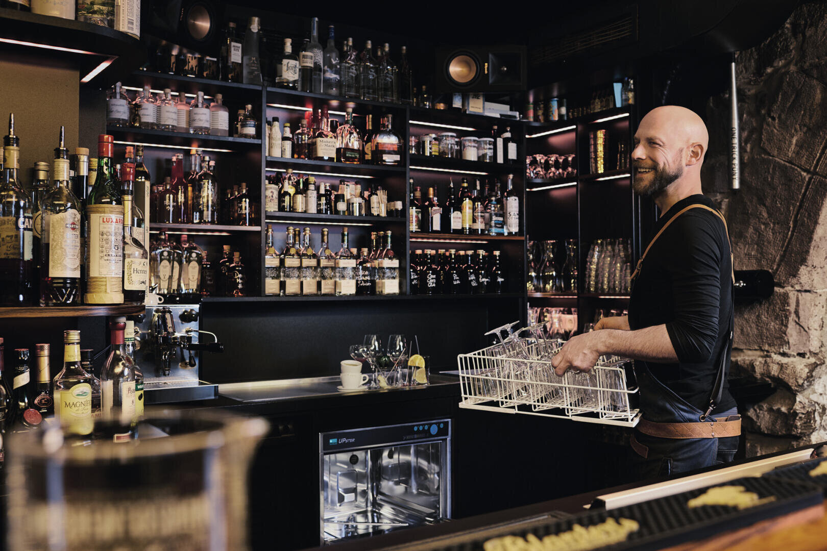 Ein Barkeeper lächelt, während er in einer modernen Bar, umgeben von Regalen voller Flaschen und Gläsern, saubere Gläser aus einer Spülmaschine der Marke MEIKO, Modell UPster U, ausräumt.