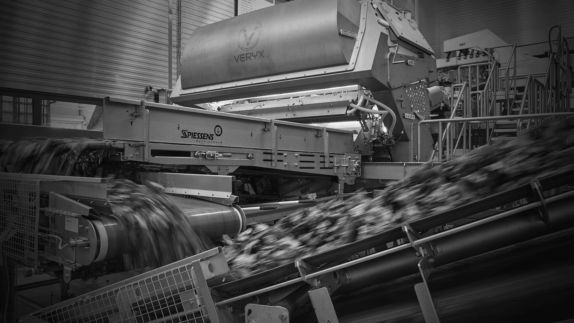 Schwarz-Weiß-Bild einer industriellen Lebensmittelverarbeitungsmaschine mit Förderbändern, die zerkleinerte Produkte zu einer großen Sortiertrommel mit der Aufschrift VERYX transportieren. Die Maschinen sind von Metallgerüsten in einer Fabrik umgeben.
