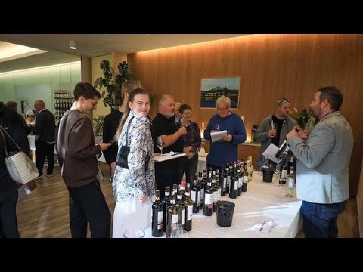 In einem hell beleuchteten Raum versammeln sich Menschen um einen Tisch mit Weinflaschen, probieren Wein und unterhalten sich darüber. Einige halten Gläser oder Notizblöcke in der Hand, und die Atmosphäre wirkt gesellig und lebhaft.