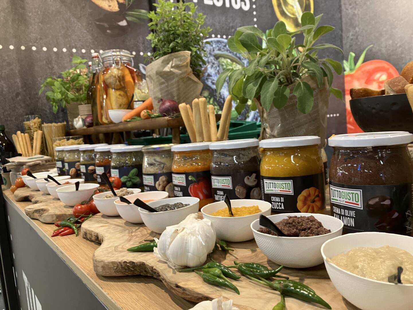 Auf einer Holztheke stehen Gläser mit Soßen und Schälchen mit Dips, die durch Grissini, frische Kräuter, Knoblauch und Chilischoten farbenfroh ergänzt werden. Im Hintergrund sind weiteres frisches Gemüse und Küchendekoration zu sehen.