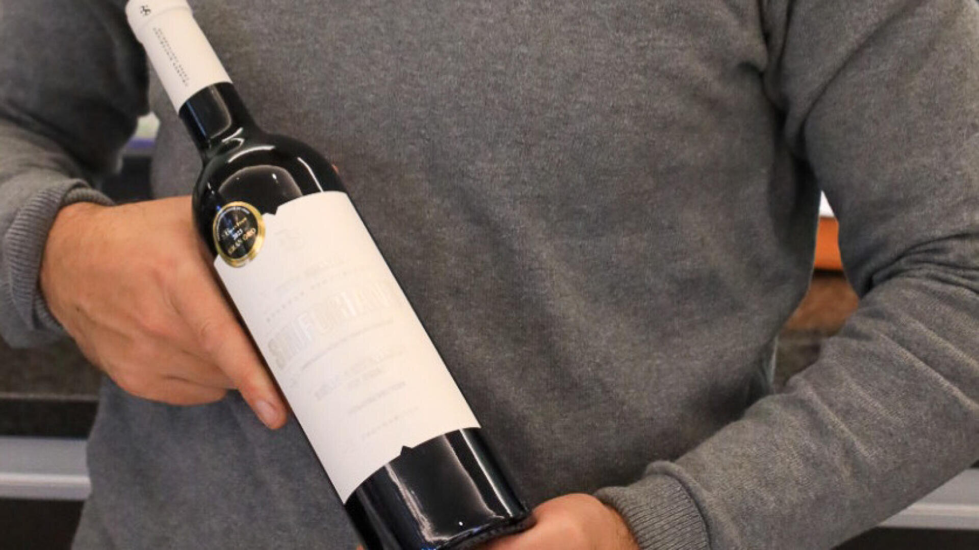 Eine Person in einem grauen Pullover hält eine Flasche Rotwein mit einem überwiegend weißen Etikett und einem goldenen Aufkleber in der Hand. Der Hintergrund scheint in einem Innenraum zu sein, möglicherweise einer Küche oder einem Barbereich.