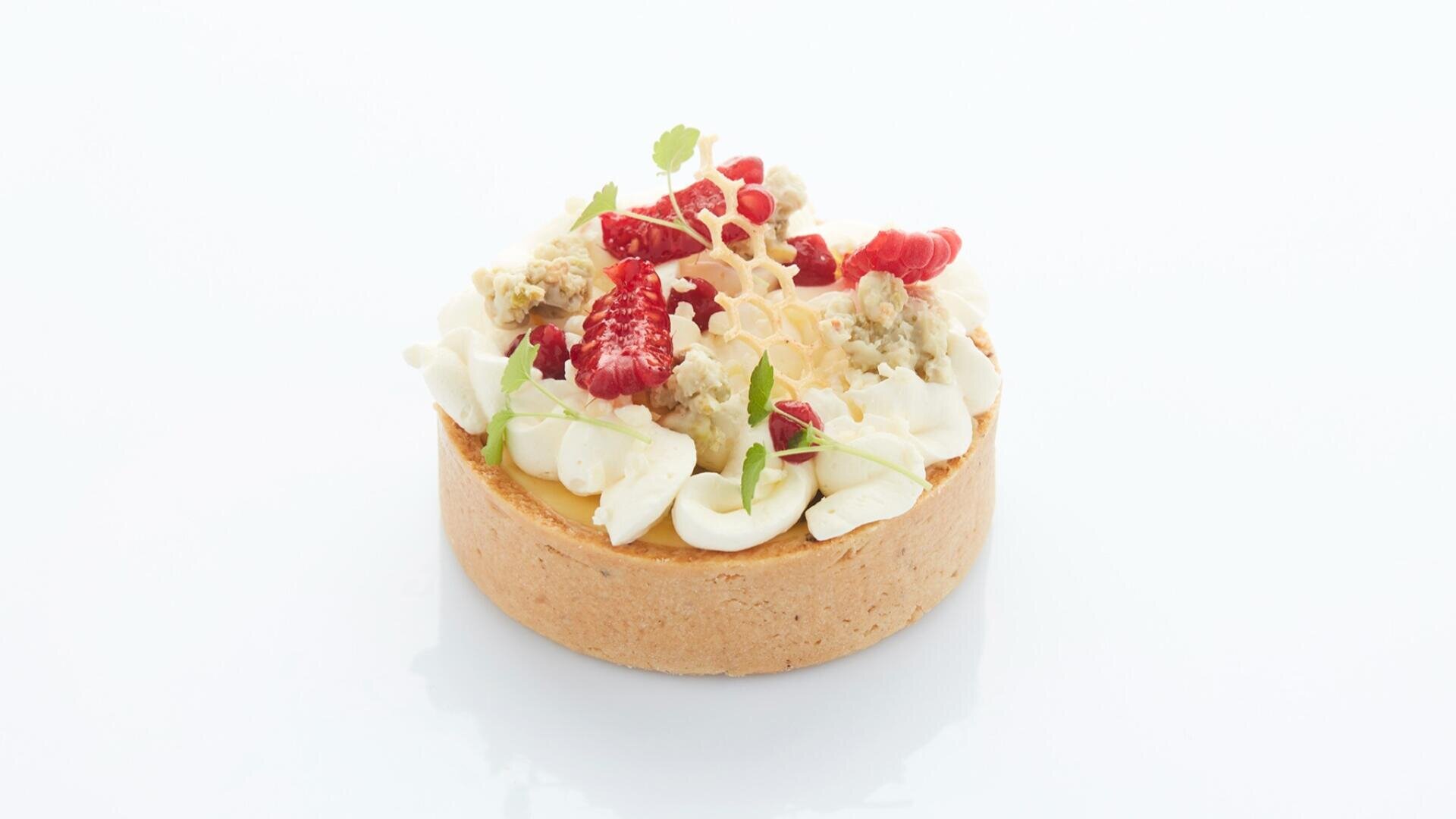 Eine runde Tarte mit goldbrauner Kruste, garniert mit weißer Sahne aus der Spritztüte, frischen Himbeeren, grünen Microgreens, Blauschimmelkäse-Krümeln und einem dekorativen Knusperbelag auf weißem Hintergrund.